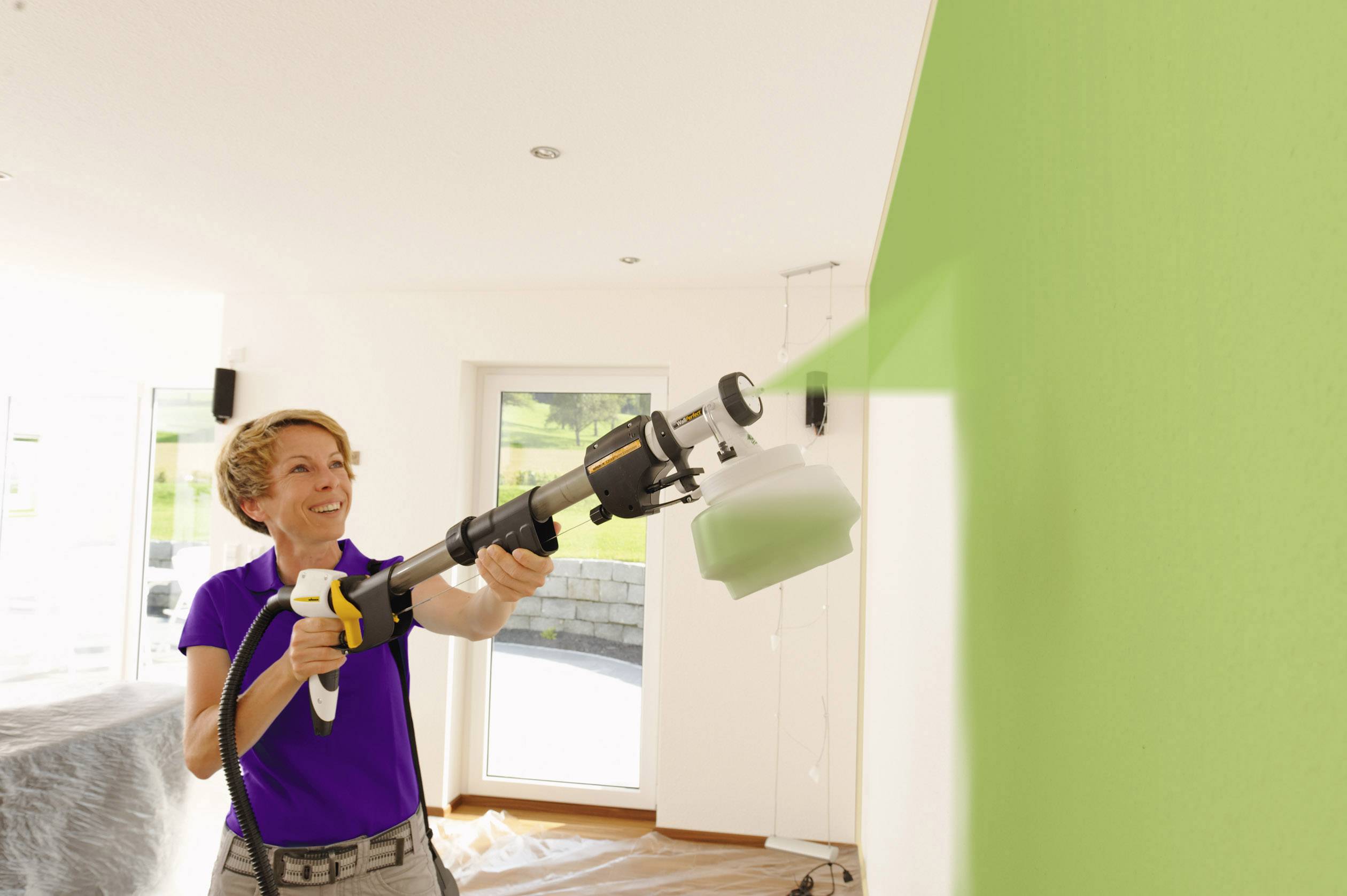 A person is spraying green paint onto an interior wall using a paint spraying system in a bright room.