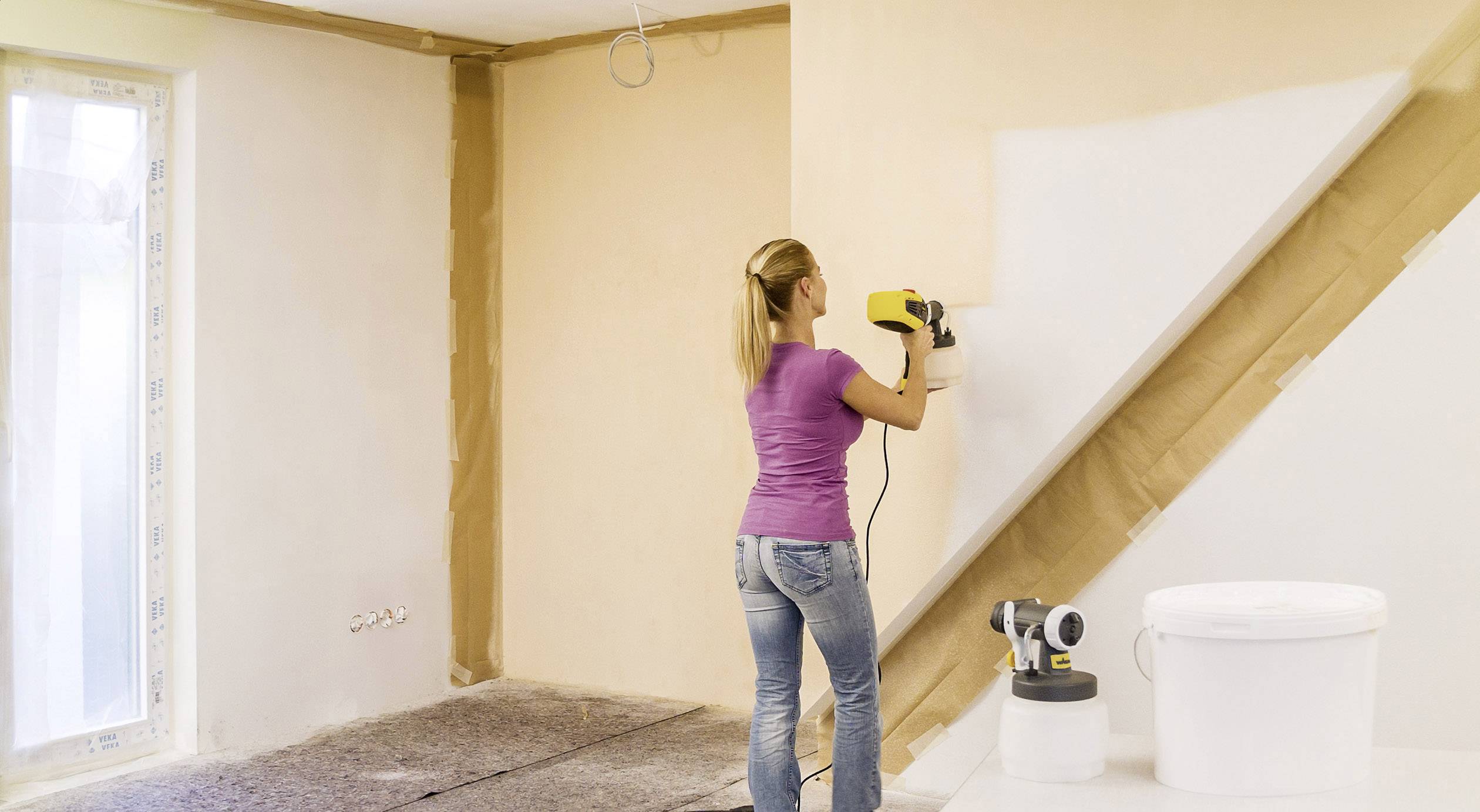 A woman is painting a wall white with a paint sprayer in a room that is being freshly renovated. Tools and a paint bucket are standing nearby.