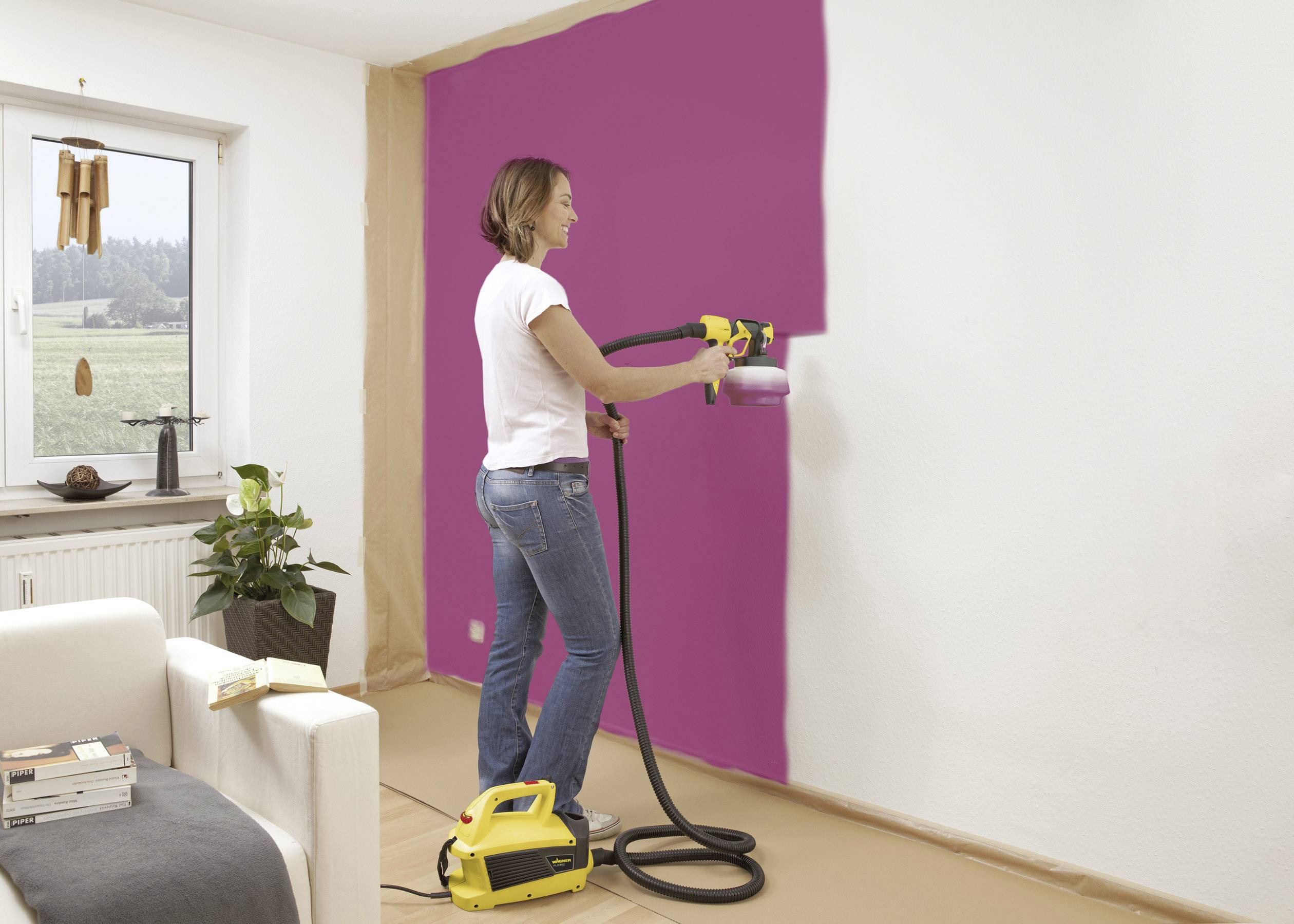A woman is using a spray paint gun to paint a white wall pink. A window with a landscape view is visible in the background.