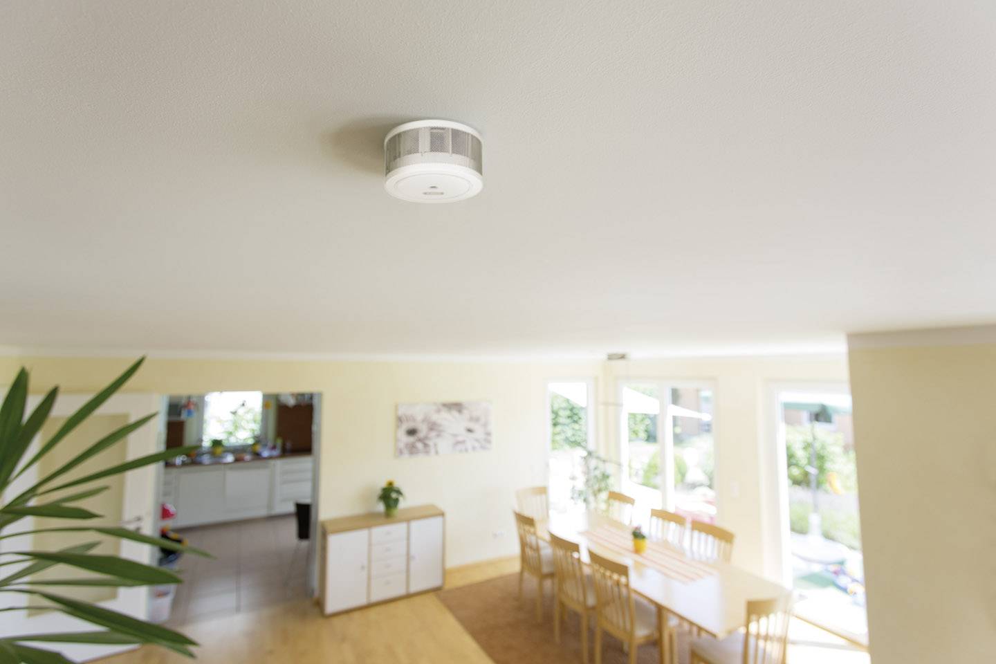A smoke detector is mounted on the ceiling of a bright living room. Below it, a table with chairs, and large windows in the background.