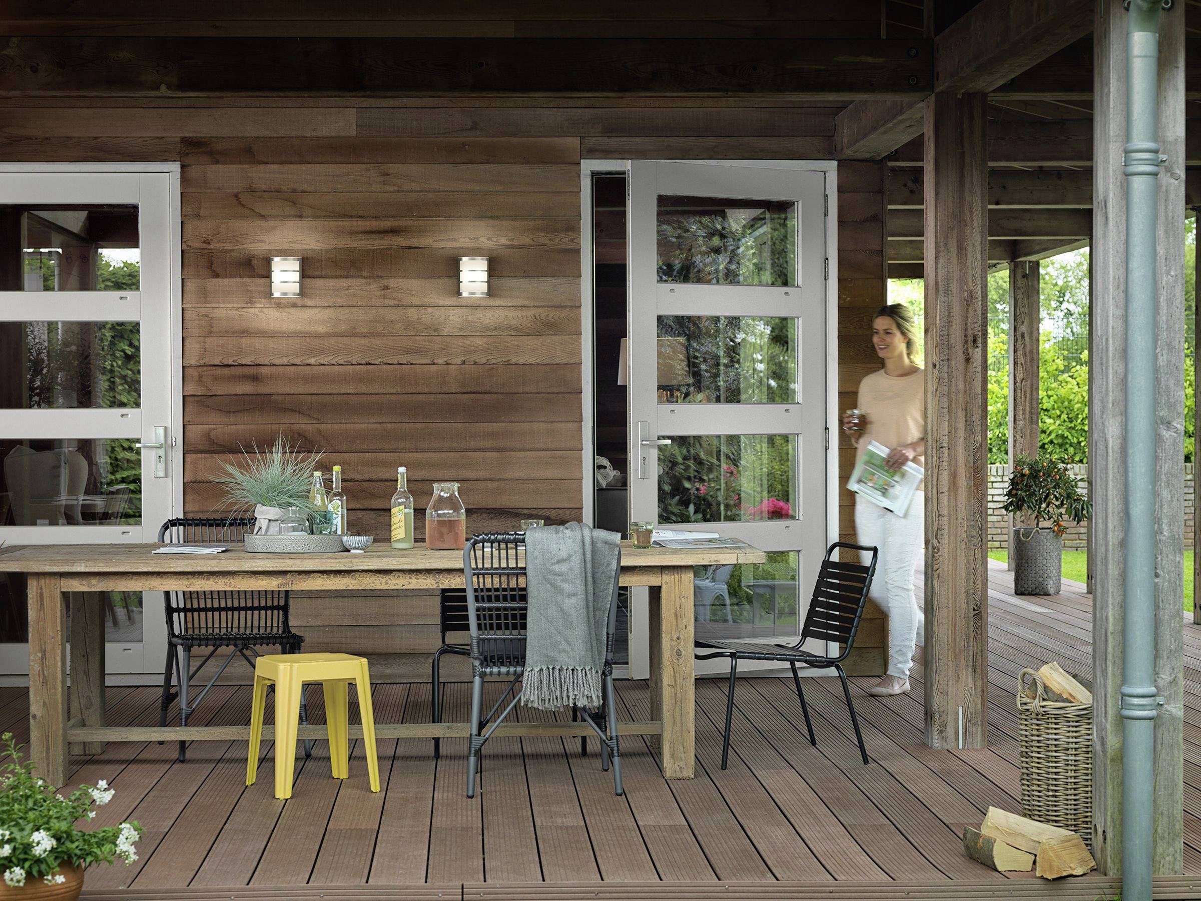 A woman enters a terrace with wooden furniture, wine bottles and glasses placed on it. A garden is visible in the background. Light falls across the scene.
