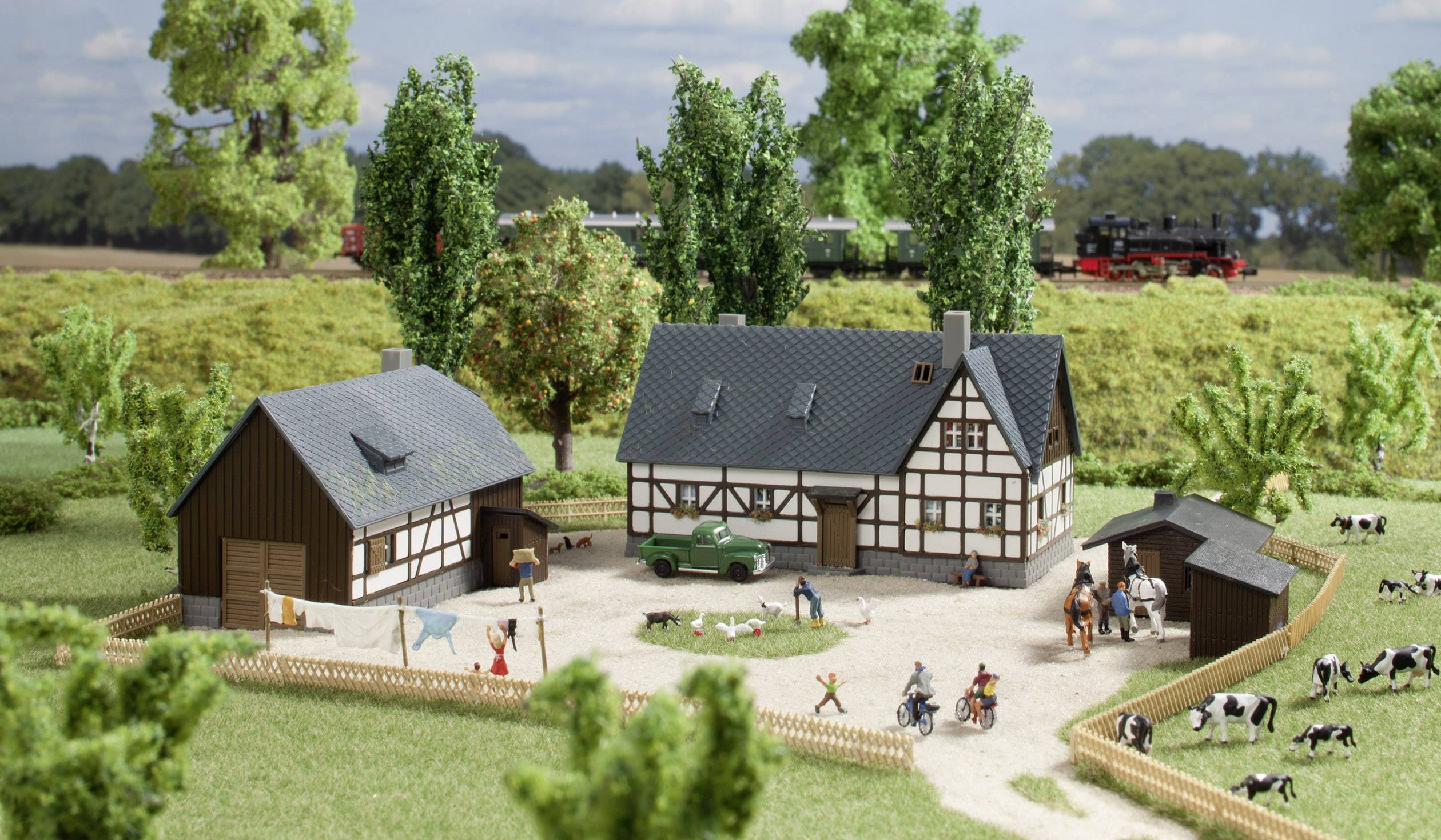 A miniature landscape depicts a farm with two half-timbered houses, surrounded by lawns and trees. Cows are grazing in the enclosed area. In the background, a train passes by.