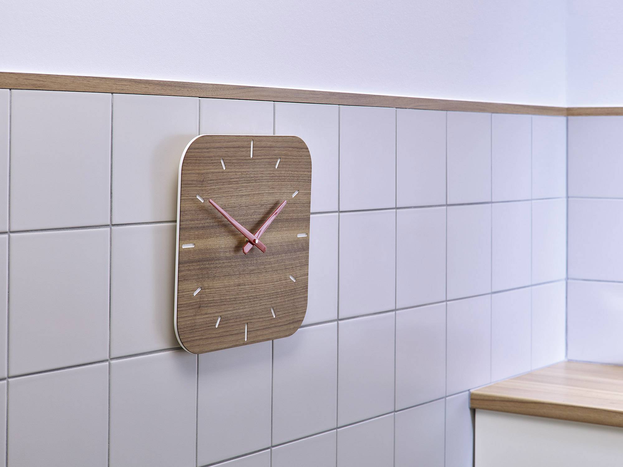 A modern wall clock with a wood-effect finish and pink hands hangs on a white-tiled wall in a kitchen.