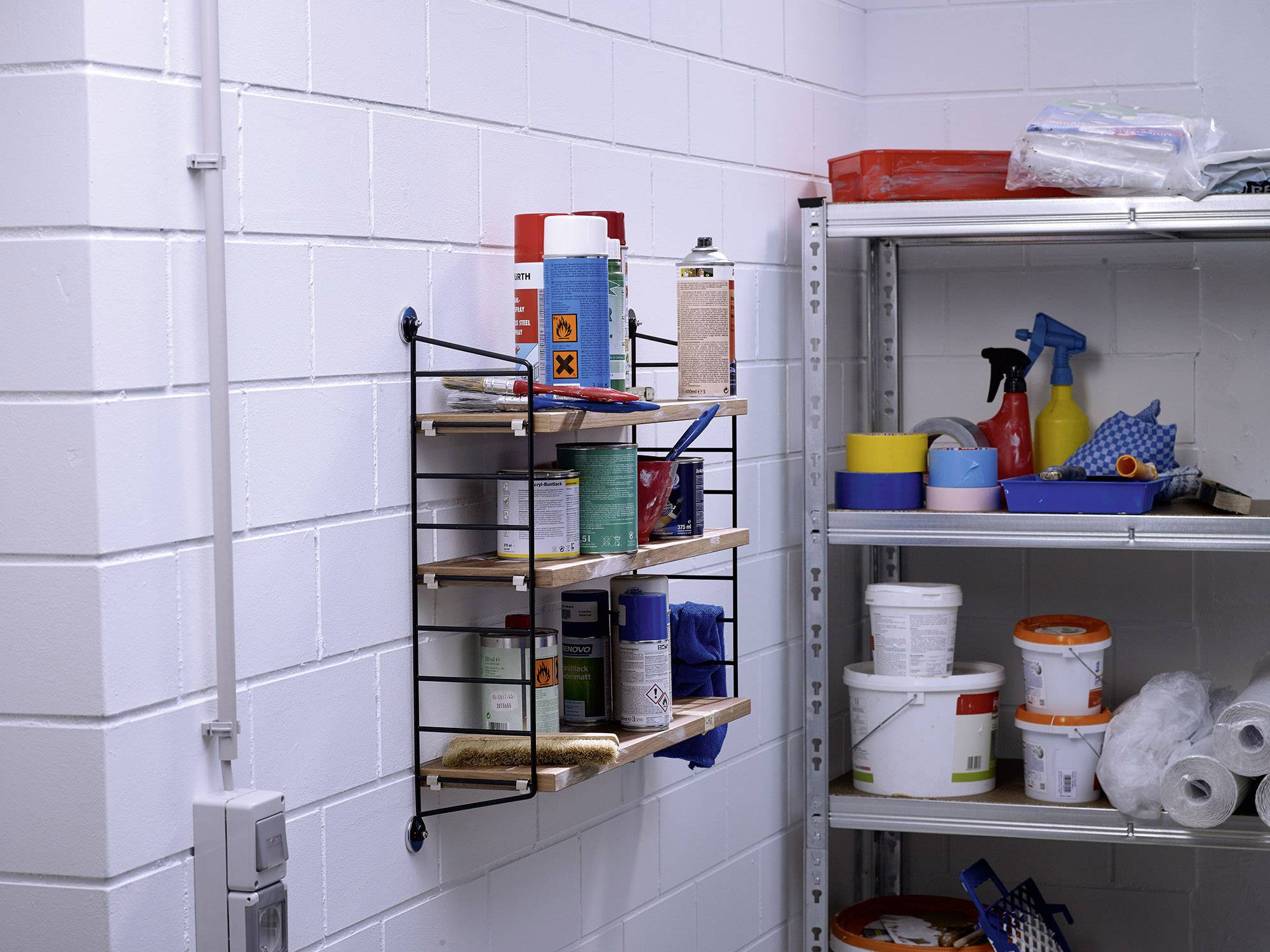 A storage cupboard with shelves against a white wall. On the left, chemical products are placed on a small wall shelf, while larger containers are positioned on a metal shelving unit to the right.