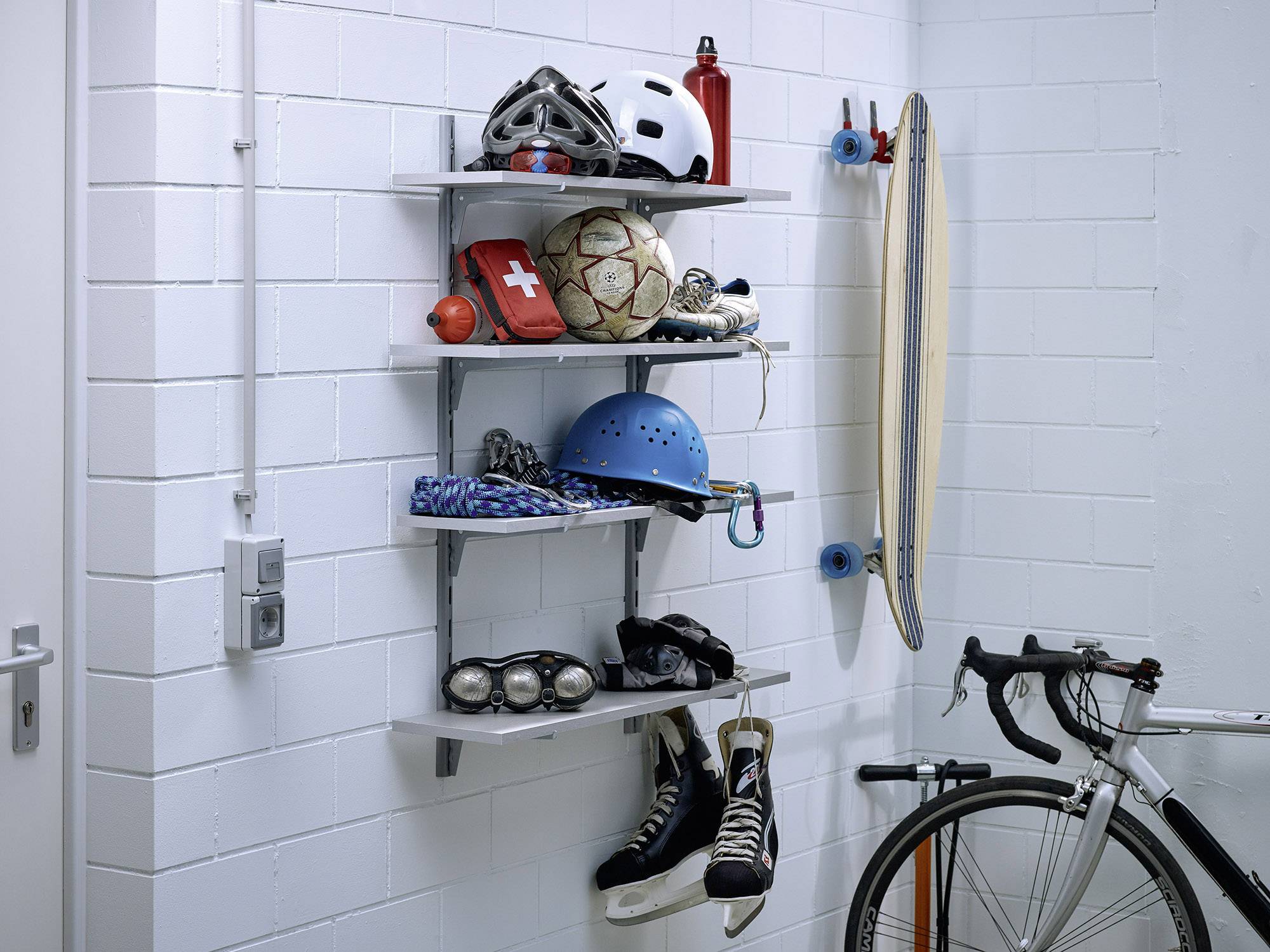 Garage wall with shelves full of sports equipment: helmets, footballs, inline skates, first aid kit; next to it a bicycle and a longboard.