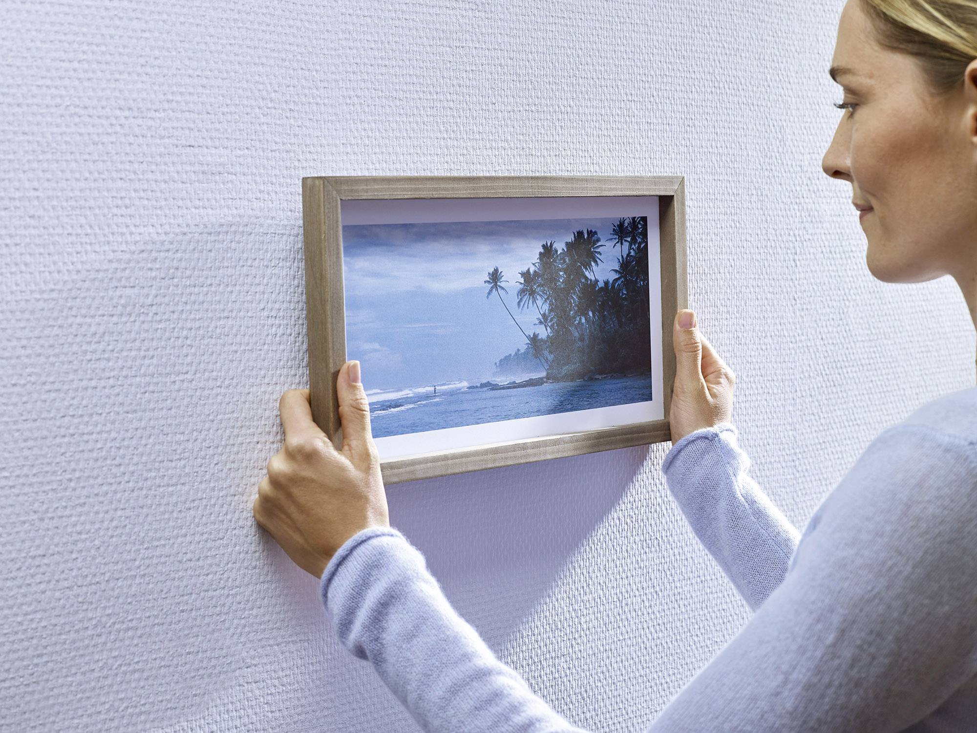A person is hanging a framed picture with a beach and palm tree scene on a white, textured wall.
