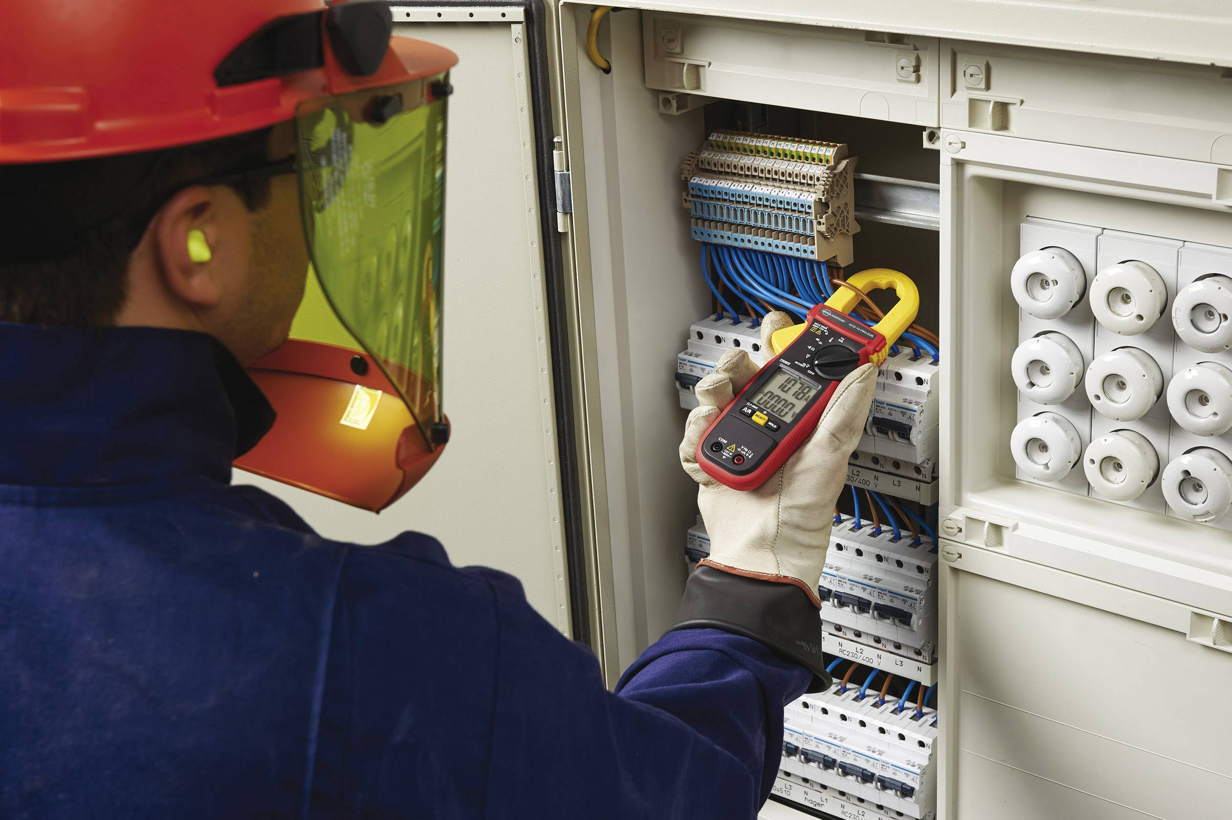 An electrician tests the voltage in an open electrical cabinet with a digital multimetre. Safety protective equipment is being worn.