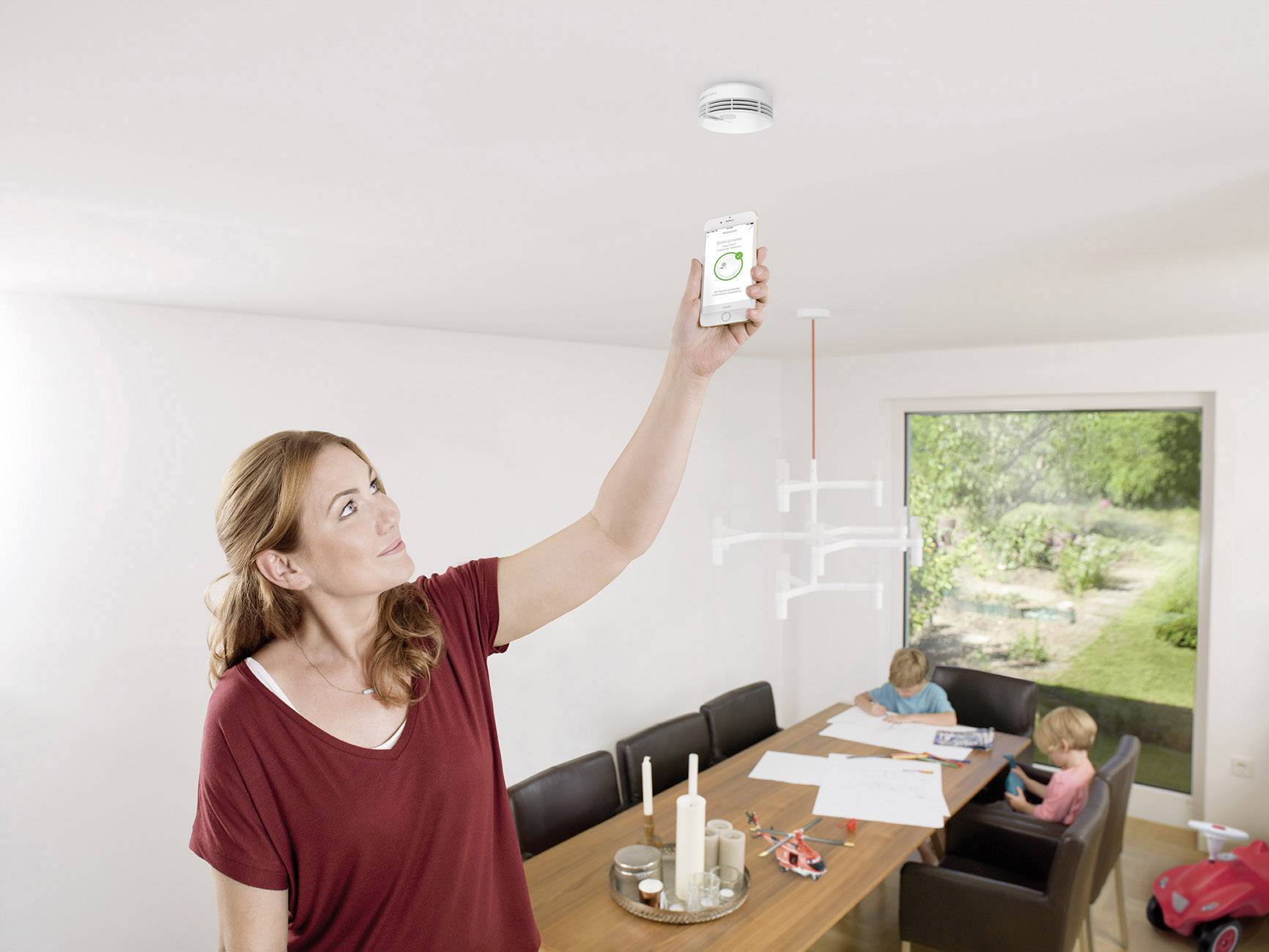 A woman checks a smoke detector with a smartphone in a brightly lit dining room, while two children are drawing at the table.