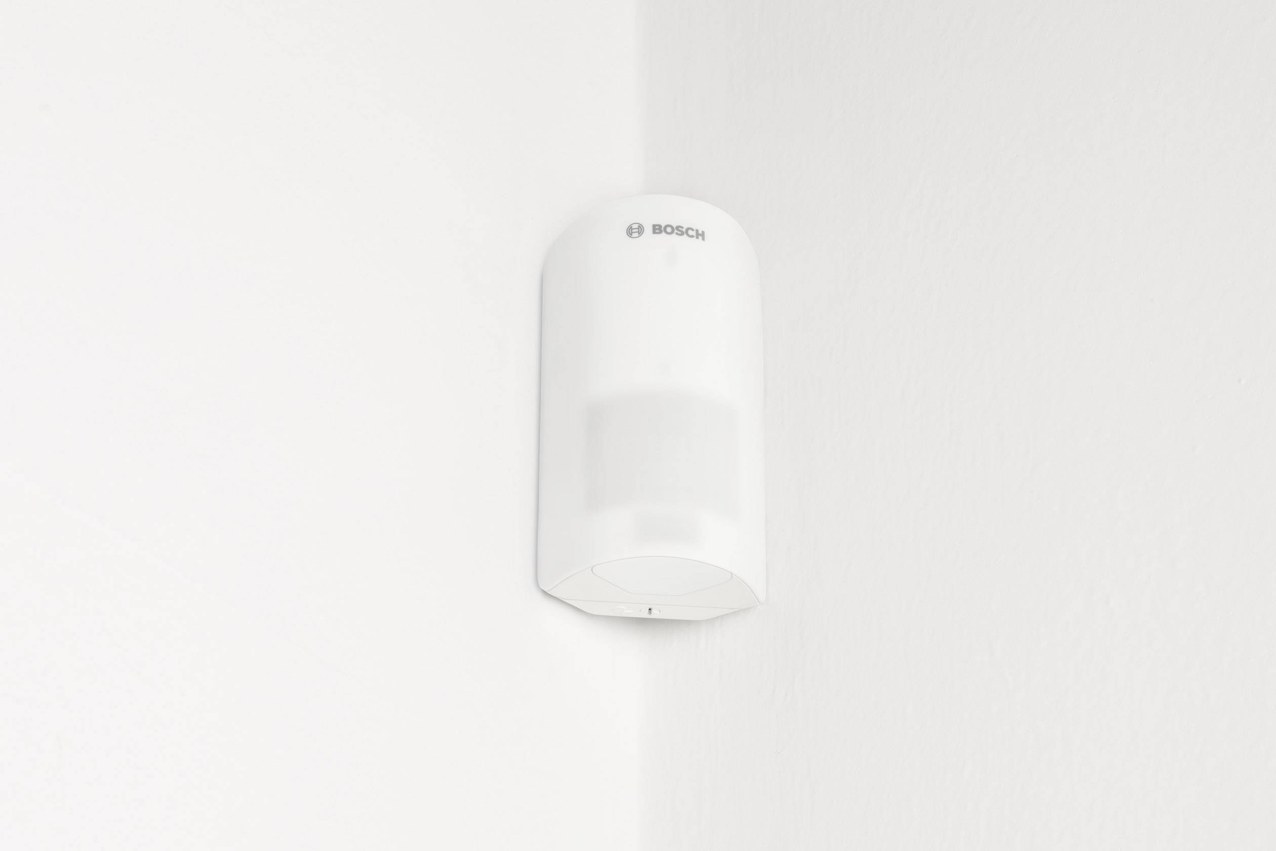 A white motion sensor is mounted on the corner of a white wall.