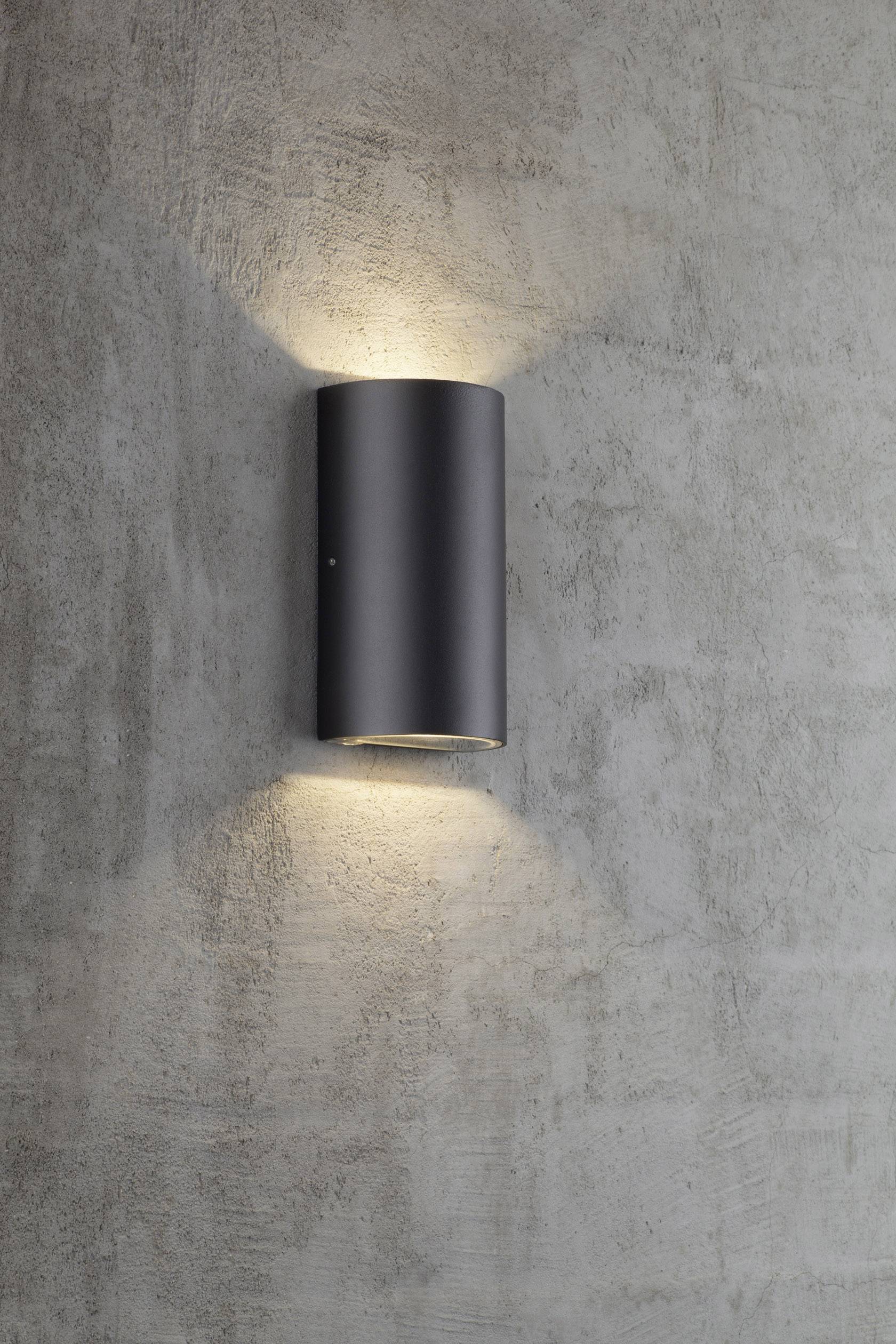 Wall light in a modern, cylindrical design, casting light upwards and downwards against a grey concrete wall.