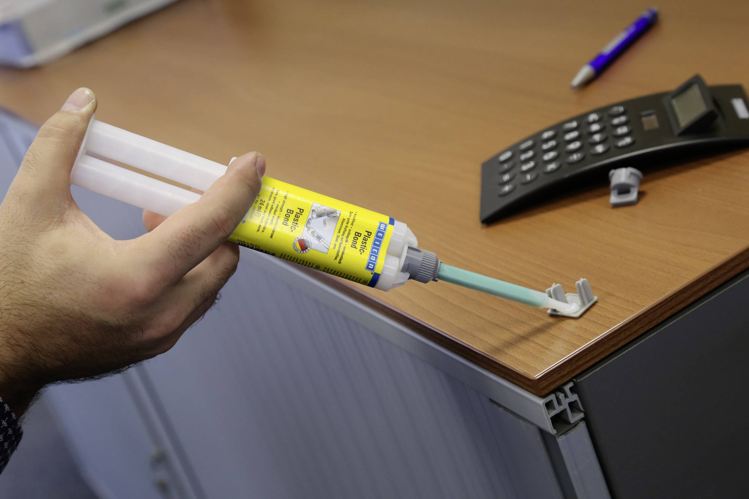 A person is applying adhesive with a double cartridge to a metal element on the edge of a table. A calculator is located on the table.