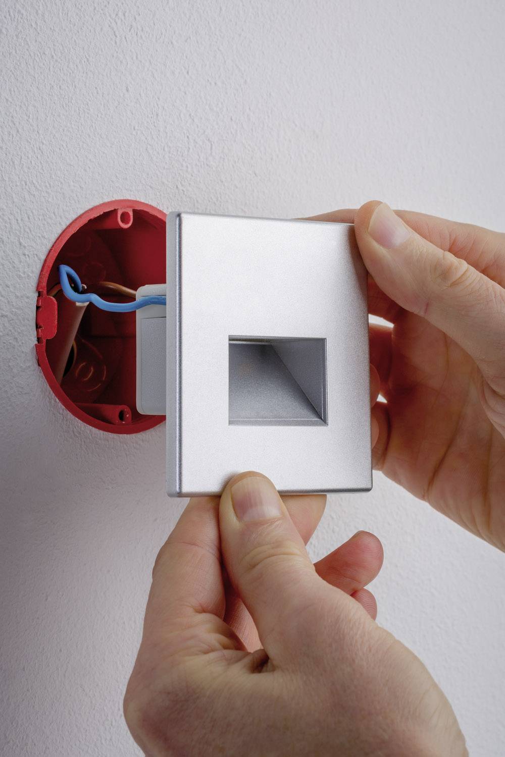 A person is installing a socket cover on an exposed electrical socket with blue wires in the wall.