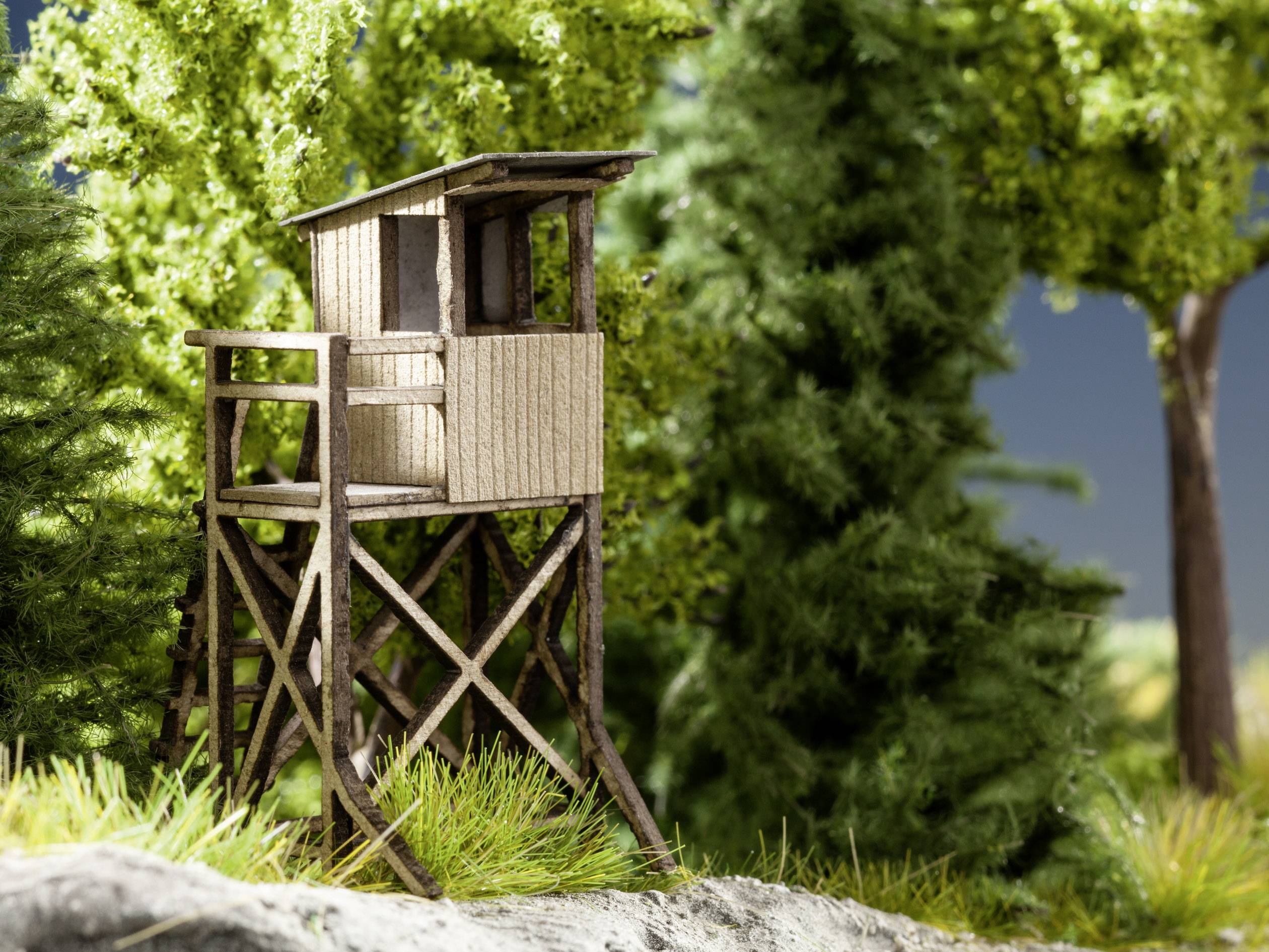 A wooden hunting high seat stands in the forest, surrounded by green trees and shrubs, against a blue sky.