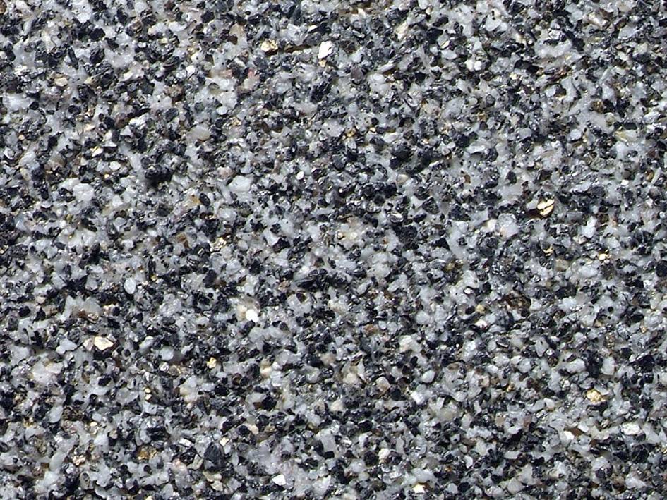 Close-up of a stony asphalt surface with irregular grey and black granulations. The texture appears coarse and rough.
