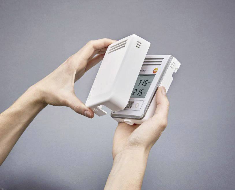A person is holding a thermostat and opening the casing to display the screen, which shows '21.5' degrees Celsius.