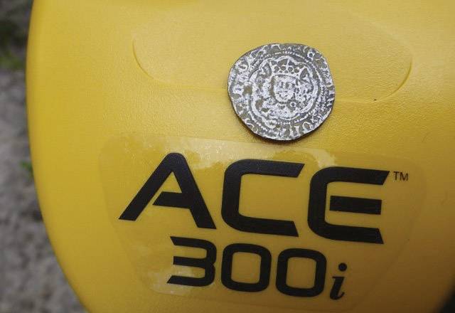 An old metal coin with an intricate pattern rests on a yellow metal detector bearing the inscription 'ACE 300i'.
