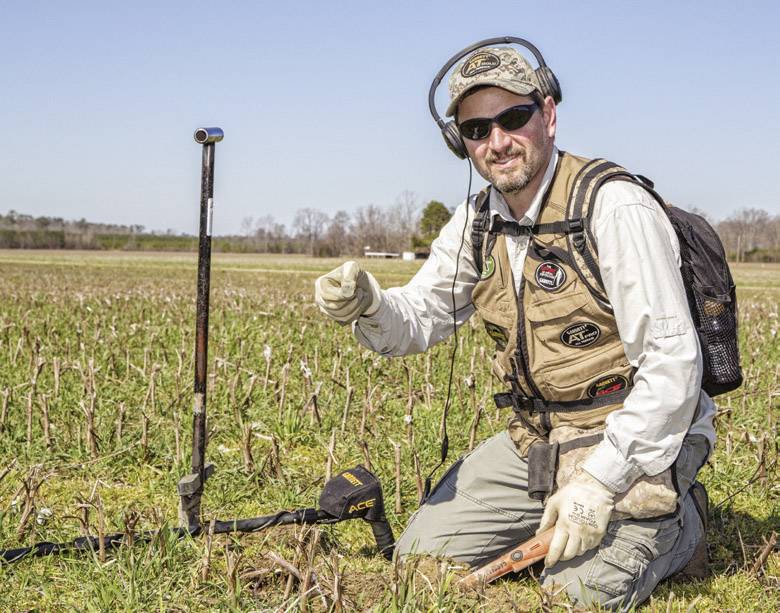 A man in casual clothing is kneeling on a meadow and holding a found object in his hand. A metal detector lies beside him.