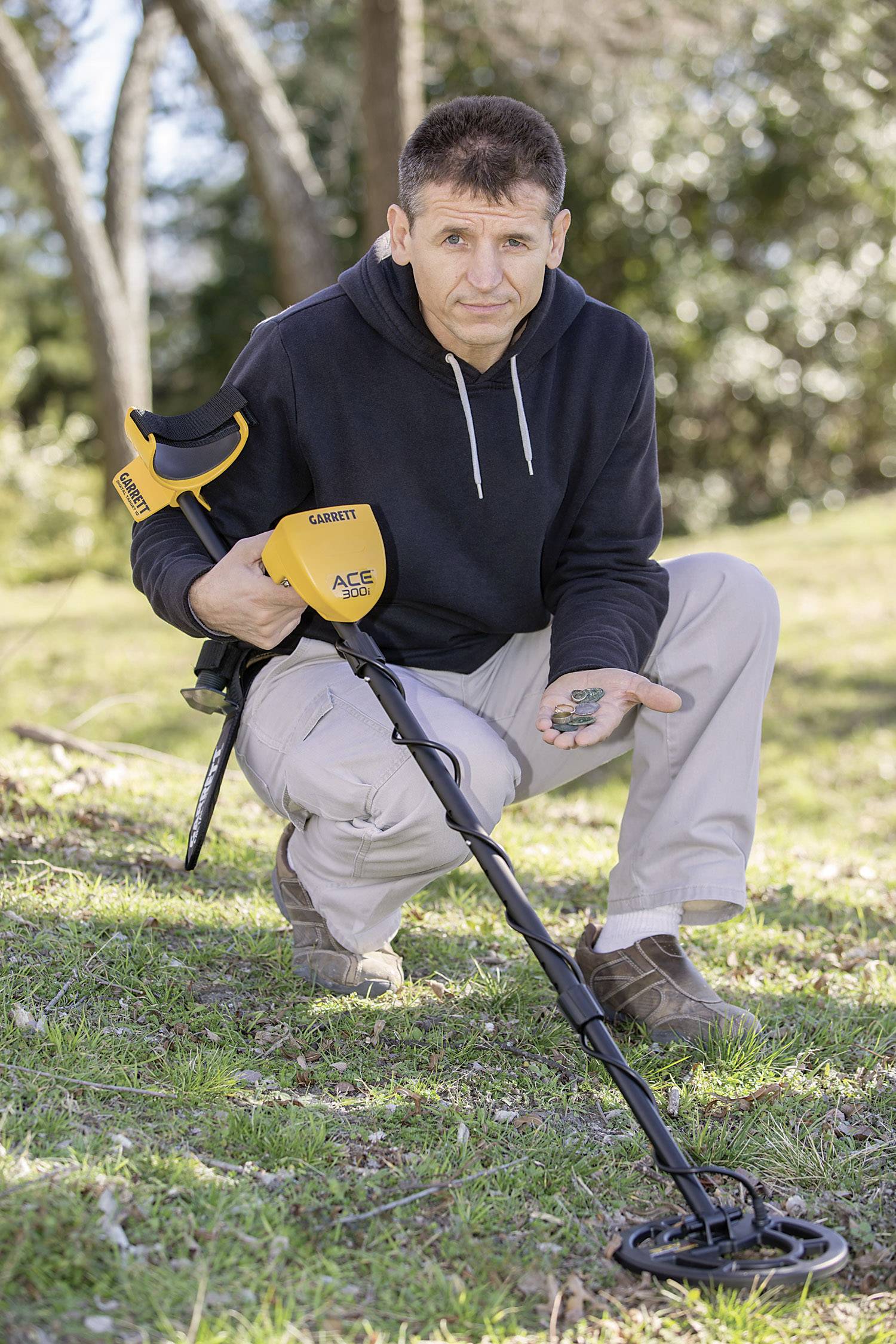 A man kneels on a grassy area, holding a yellow metal detector and proudly displaying his finds in his hand.
