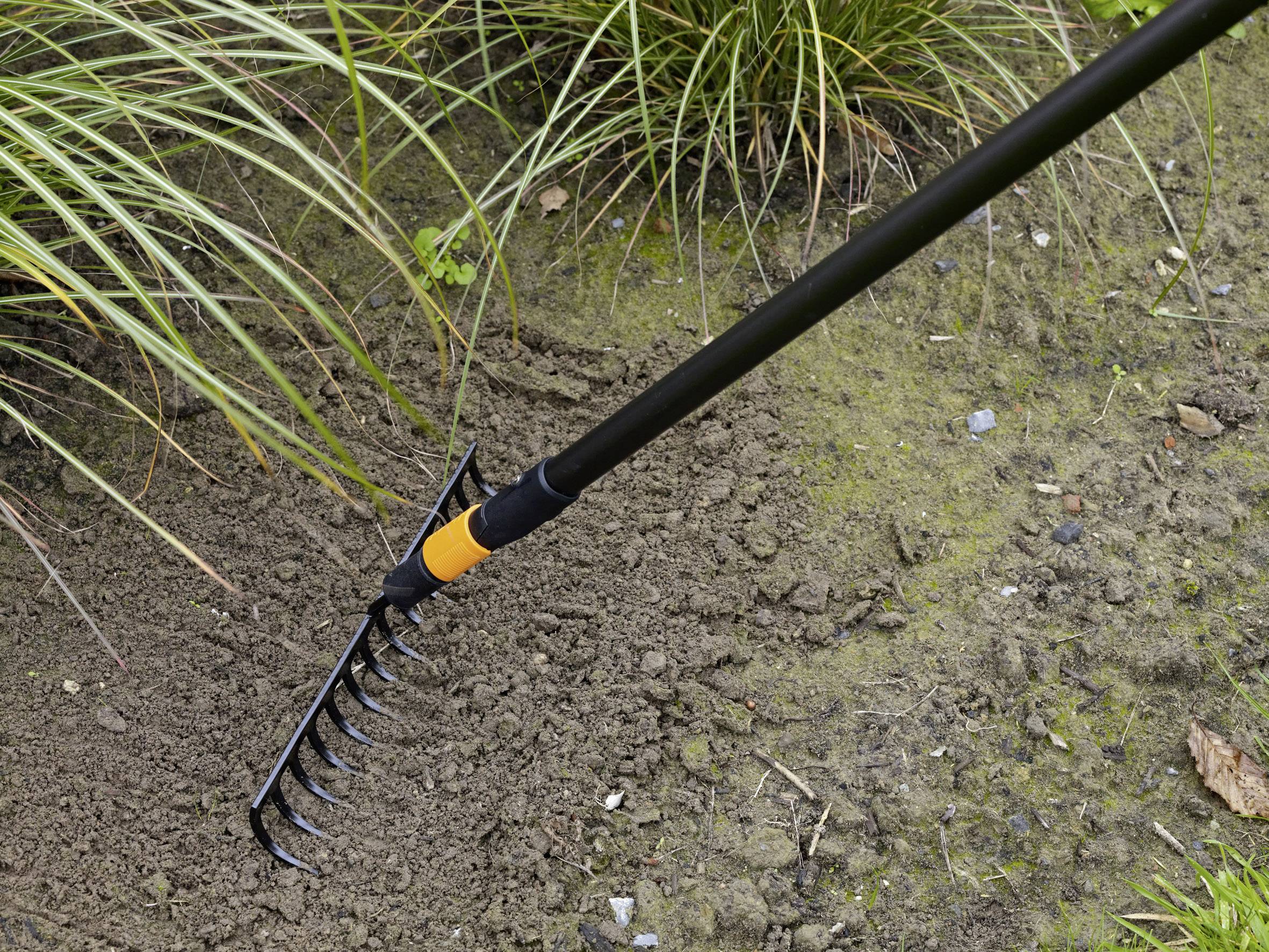 A rake is drawn over earthy ground to loosen the soil and remove weeds. In the background, tufts of grass can be seen.