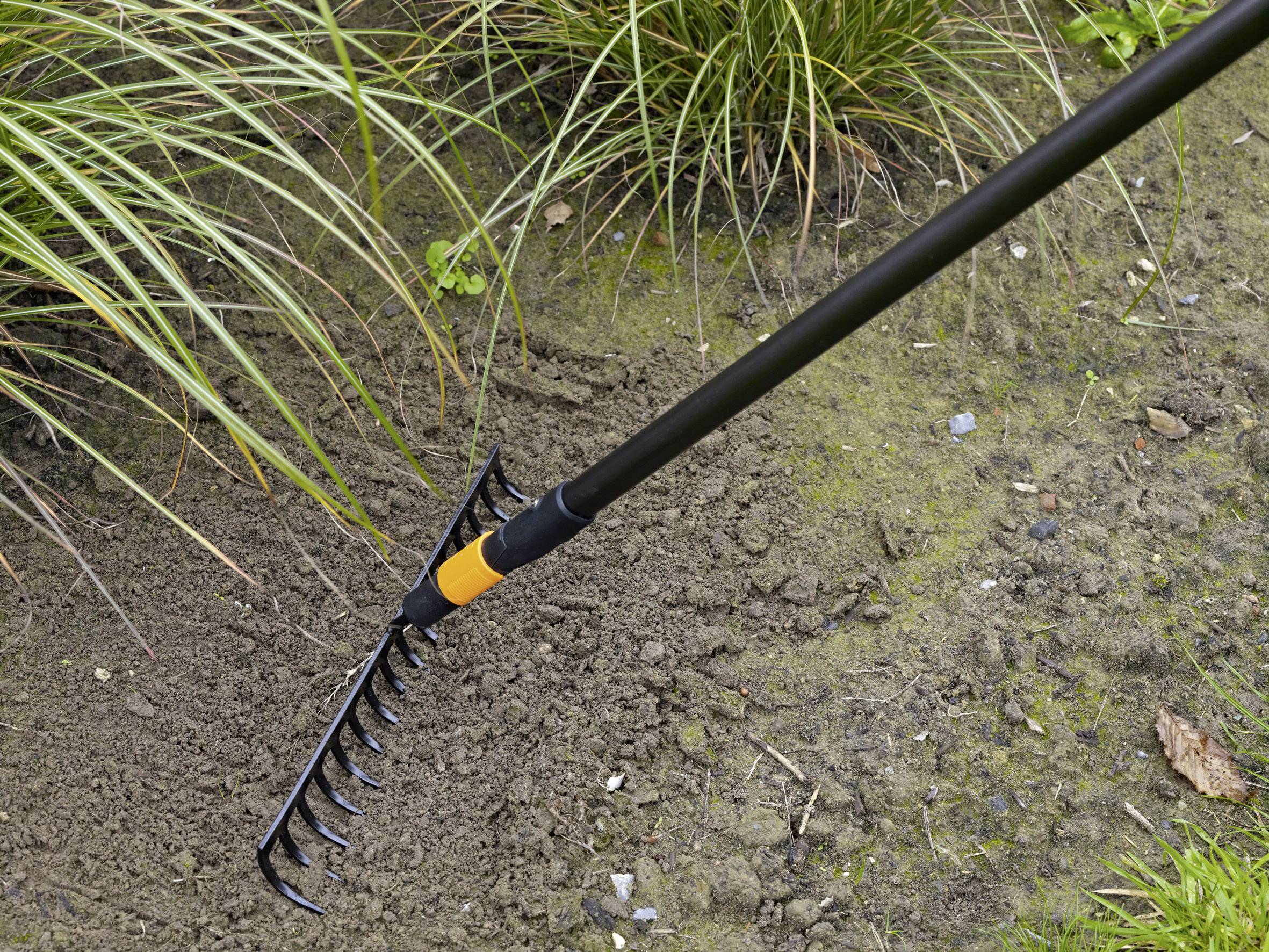 A rake is working the soil in a garden, surrounded by grass and plants, demonstrating gardening activity.