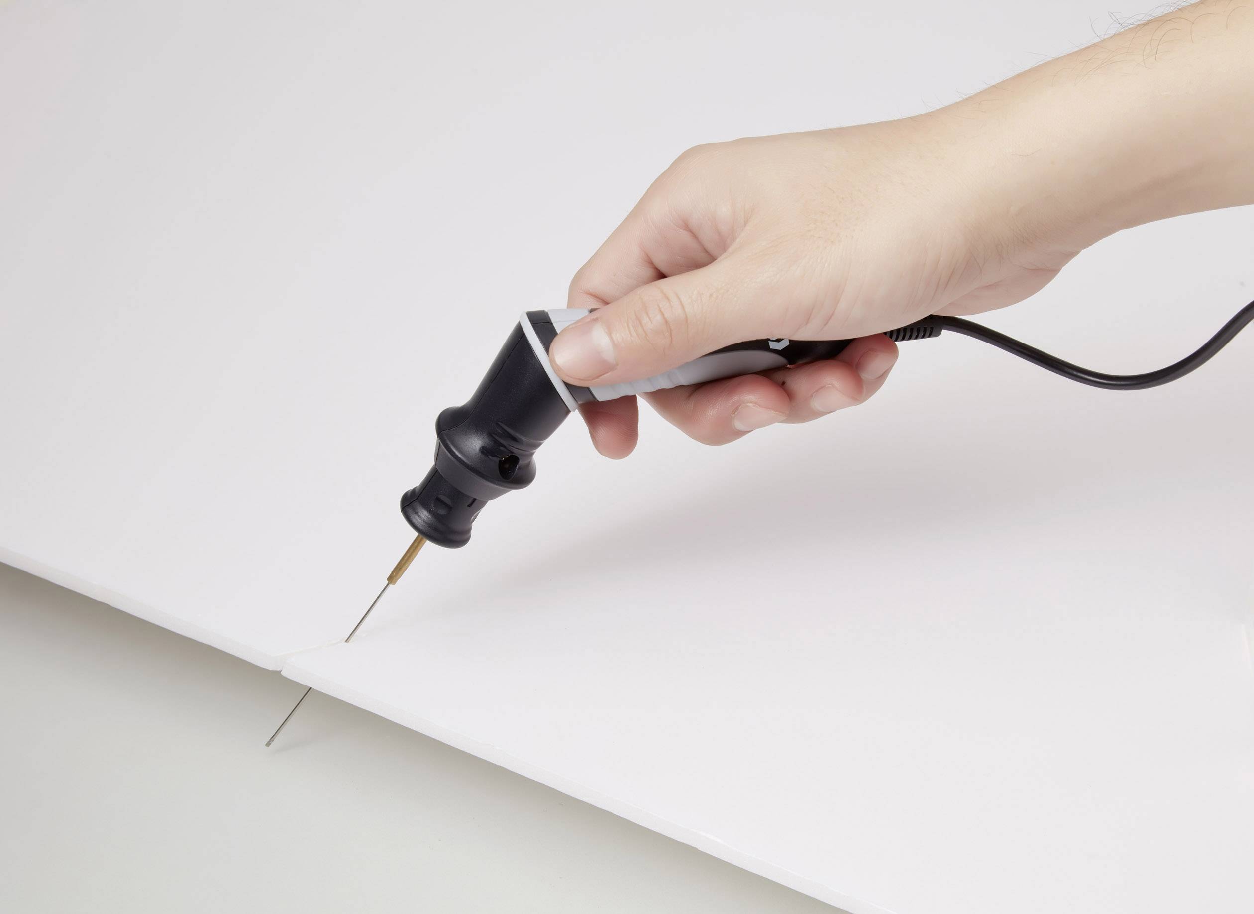 A hand is holding an electric tool with a fine tip, which is cutting into a white material.