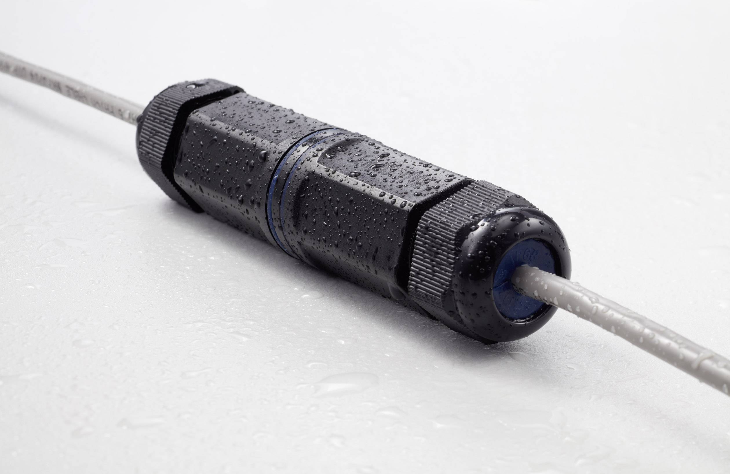 A black waterproof cable connector with water droplets on it, connected to grey cables on both sides, is resting on a smooth surface.