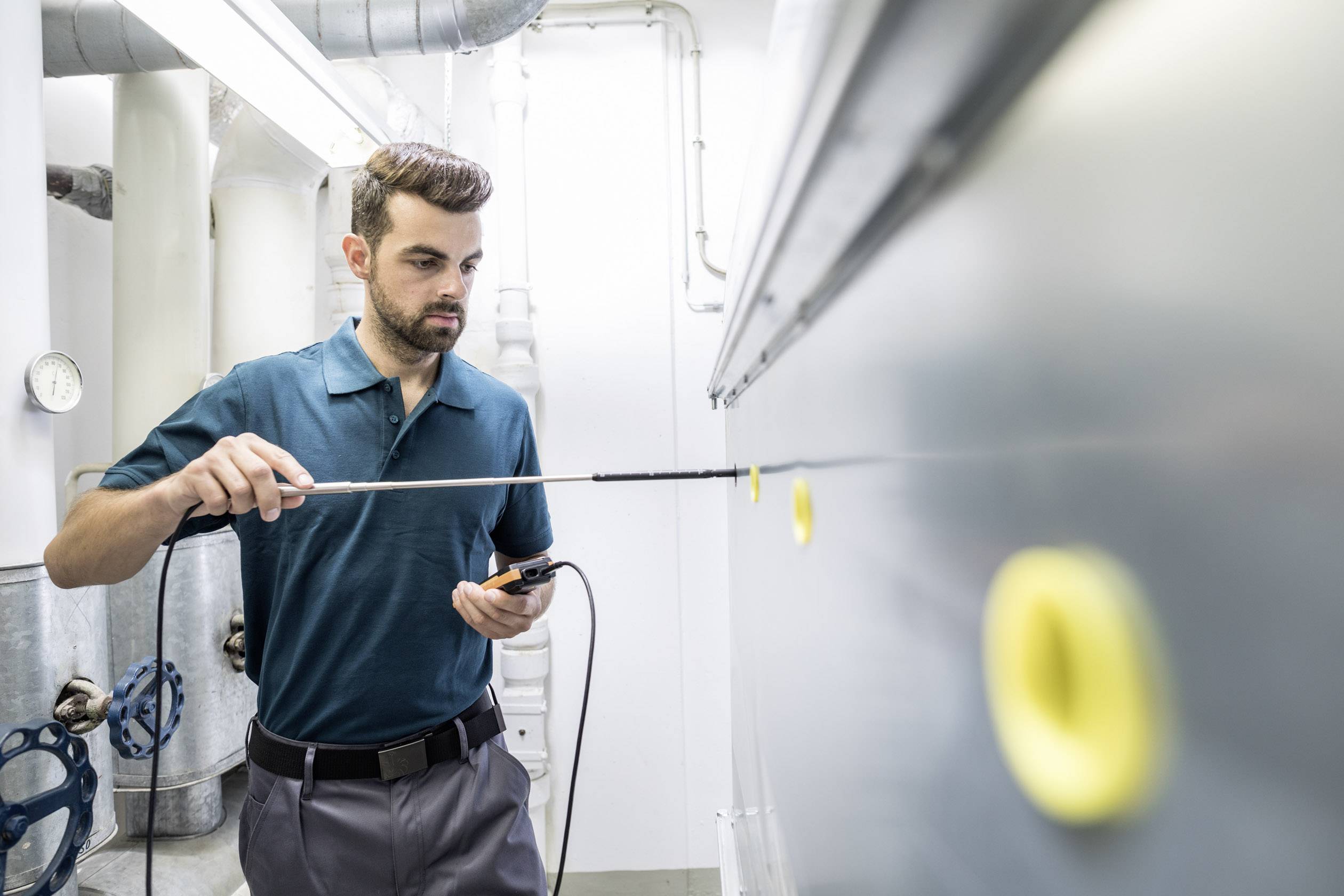 A man is inspecting an industrial pipeline with a long measuring device in a technical room. He is wearing a dark green shirt.