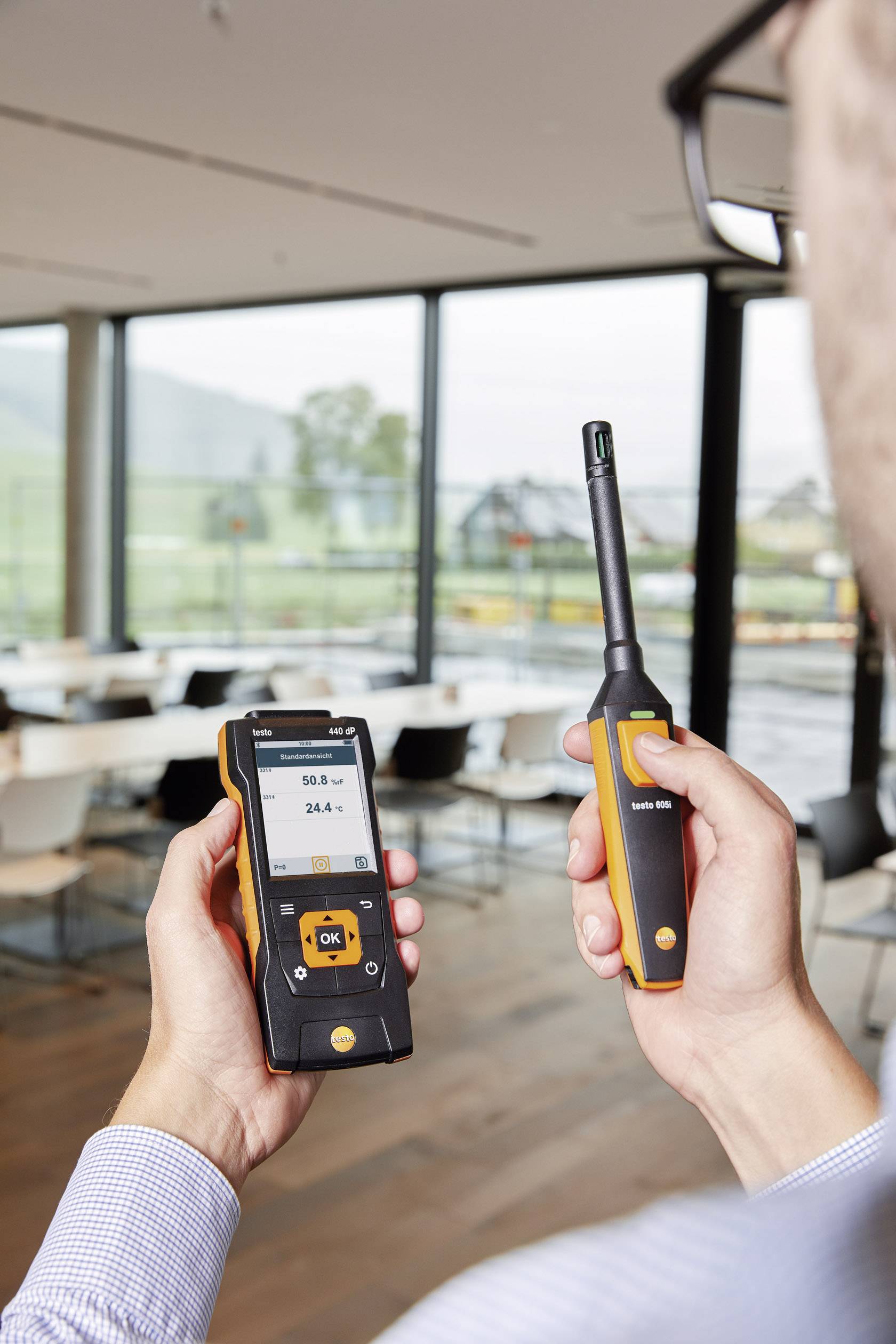 A person is holding a digital measuring device and a probe in a modern interior; the device is displaying air quality data.