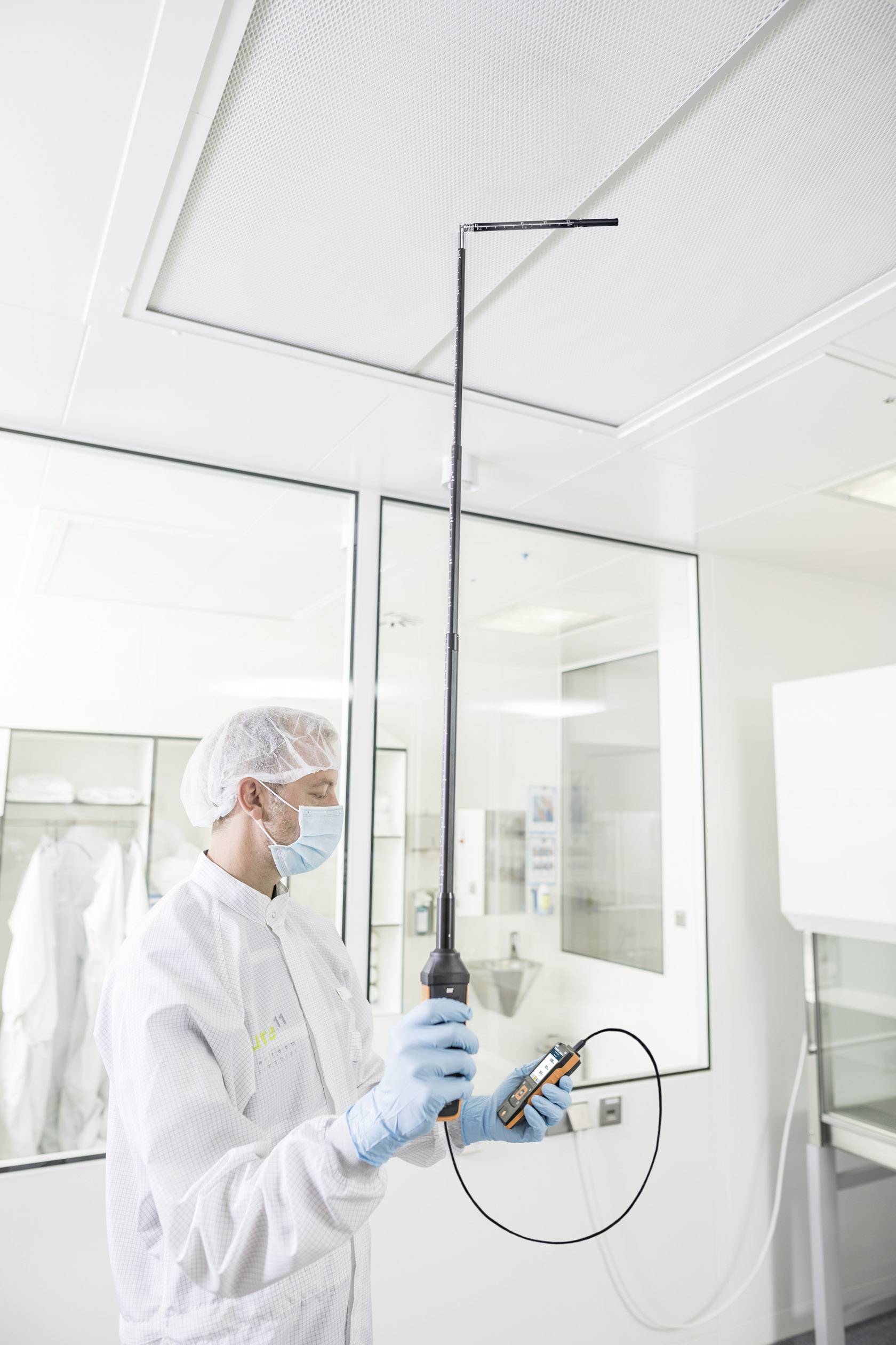 A technician in protective clothing is conducting a ventilation inspection in a clean room using a measuring instrument.
