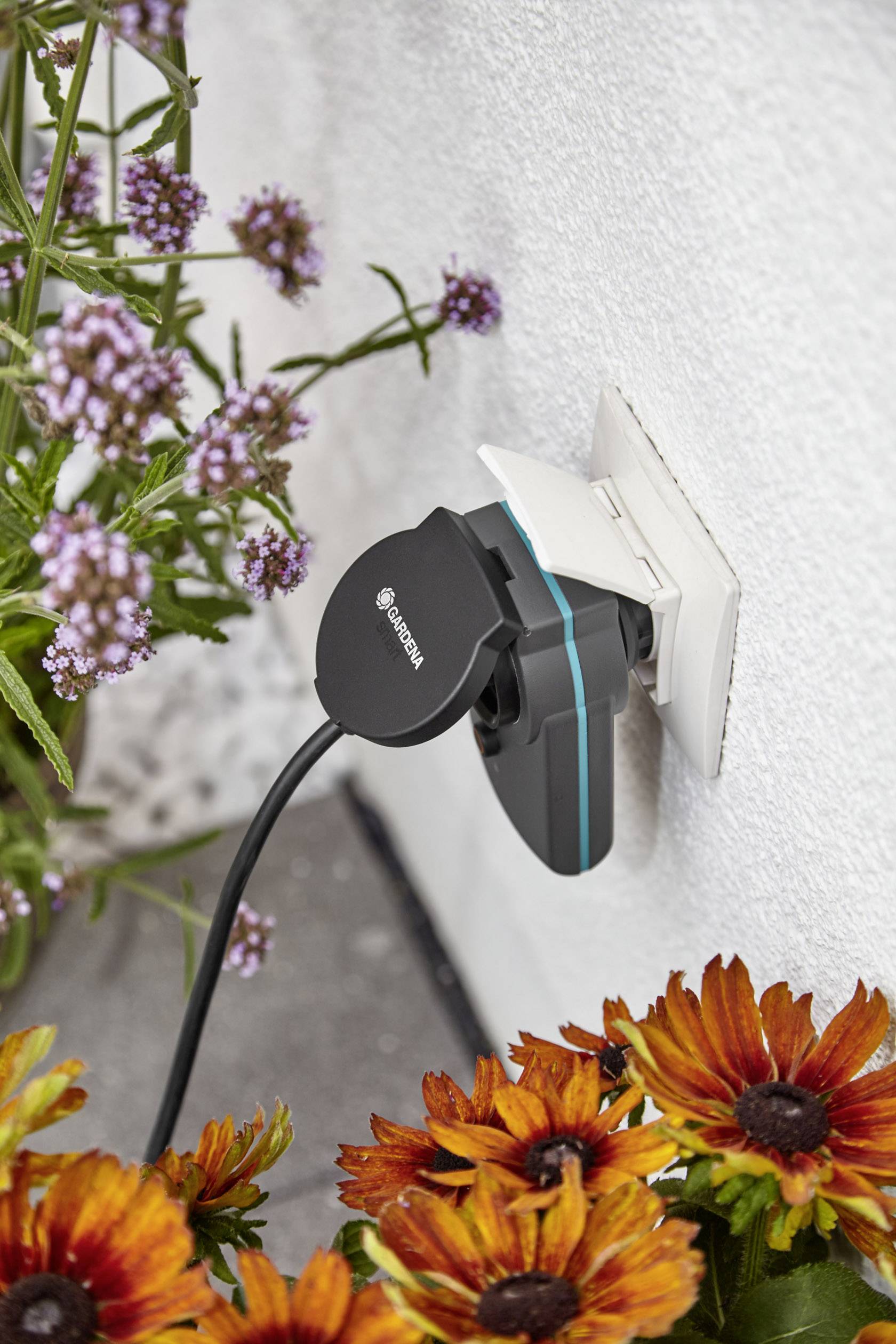 An electrical plug for outdoor use is plugged into a wall socket, surrounded by blooming flowers.