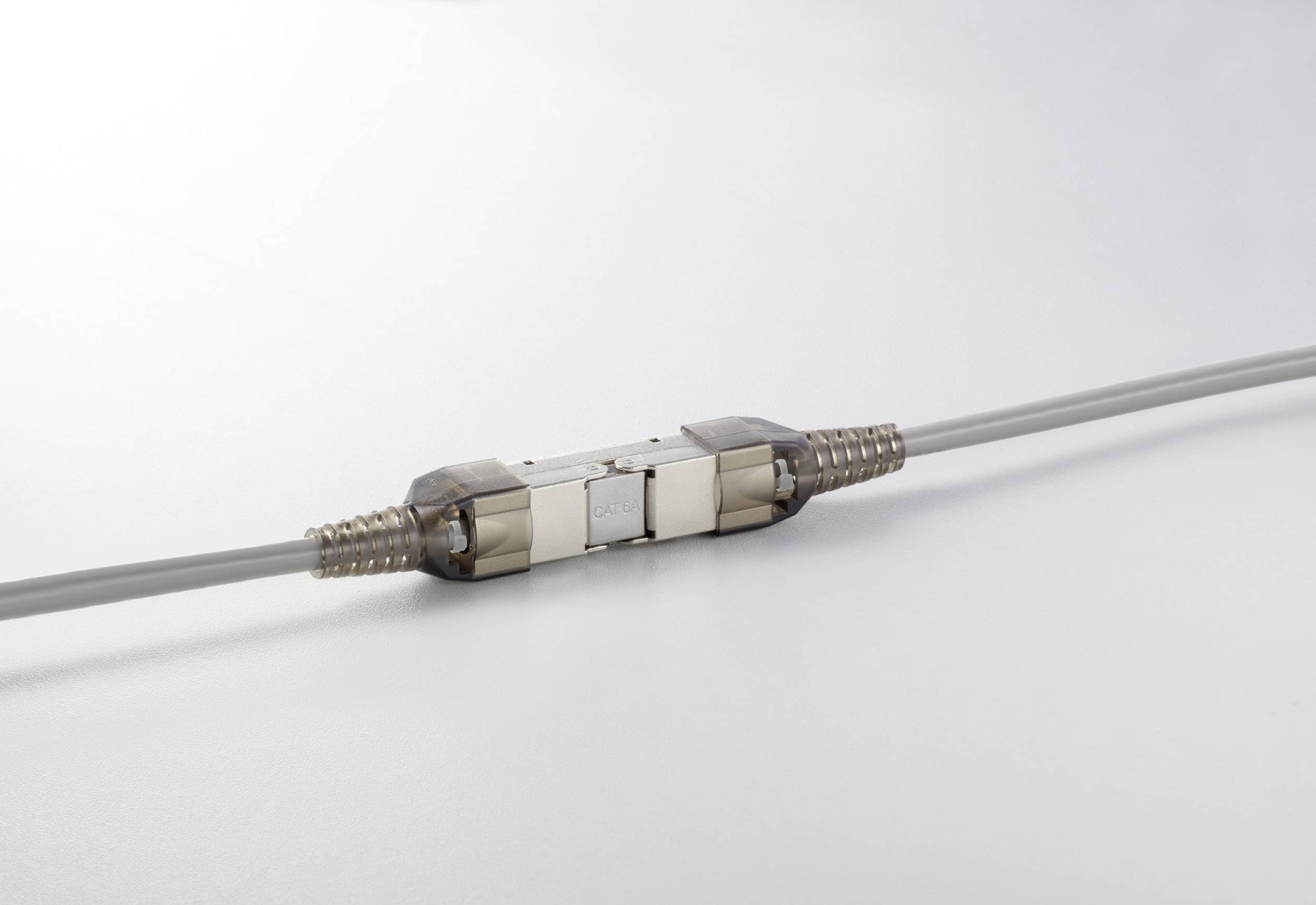 A grey cable with a metallic connector in the middle lies on a white surface. The connector has a rectangular shape.