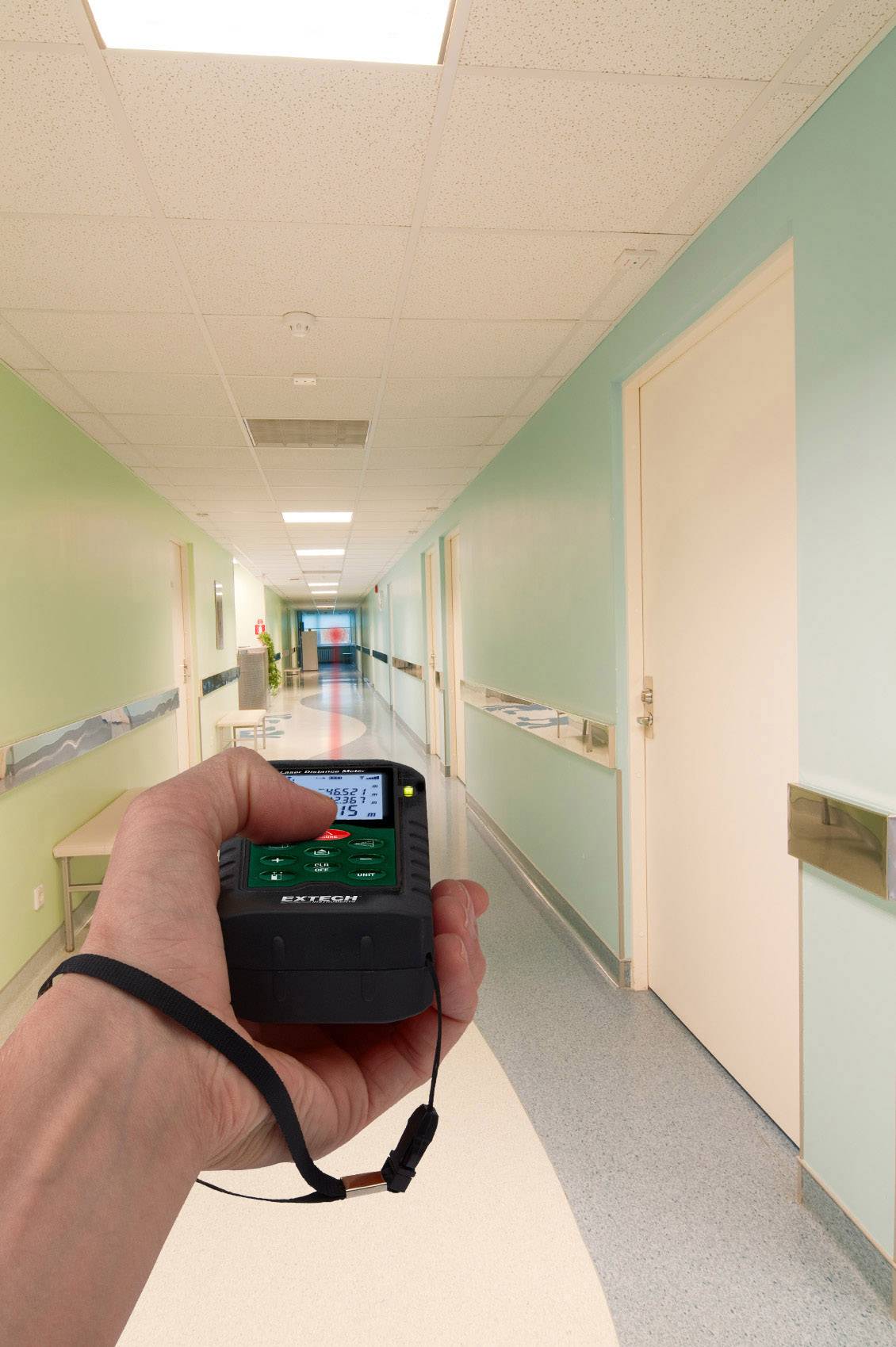 A hand is holding a laser distance measuring device in a long, brightly lit corridor with green walls and closed doors.