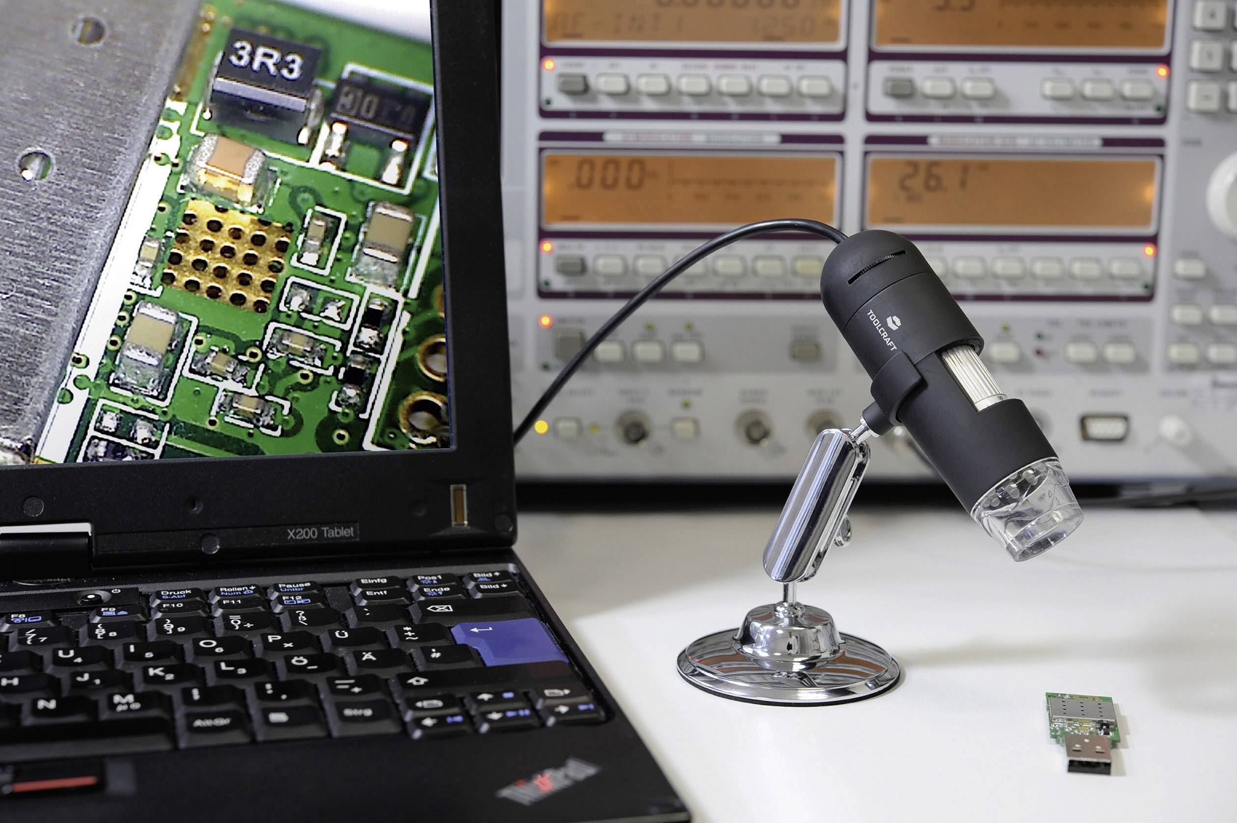 A microscope with a camera is focused on an electronic circuit board, which is magnified and displayed on a laptop screen. Measuring instruments are visible in the background.