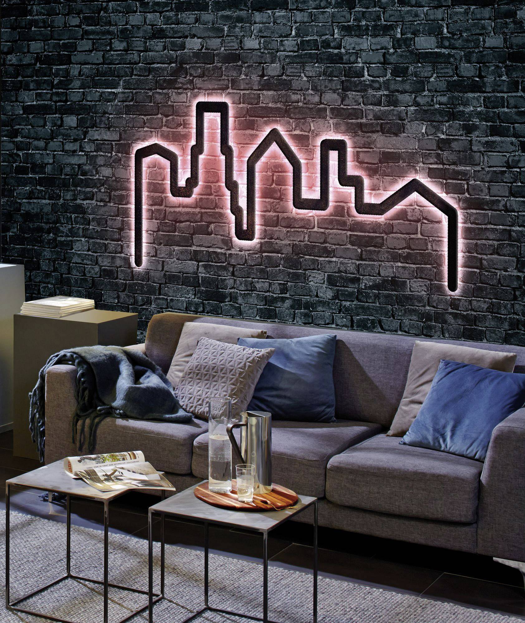 Neon cityscape of a skyline on a brick wall behind a grey sofa with cushions, with two small coffee tables in front.