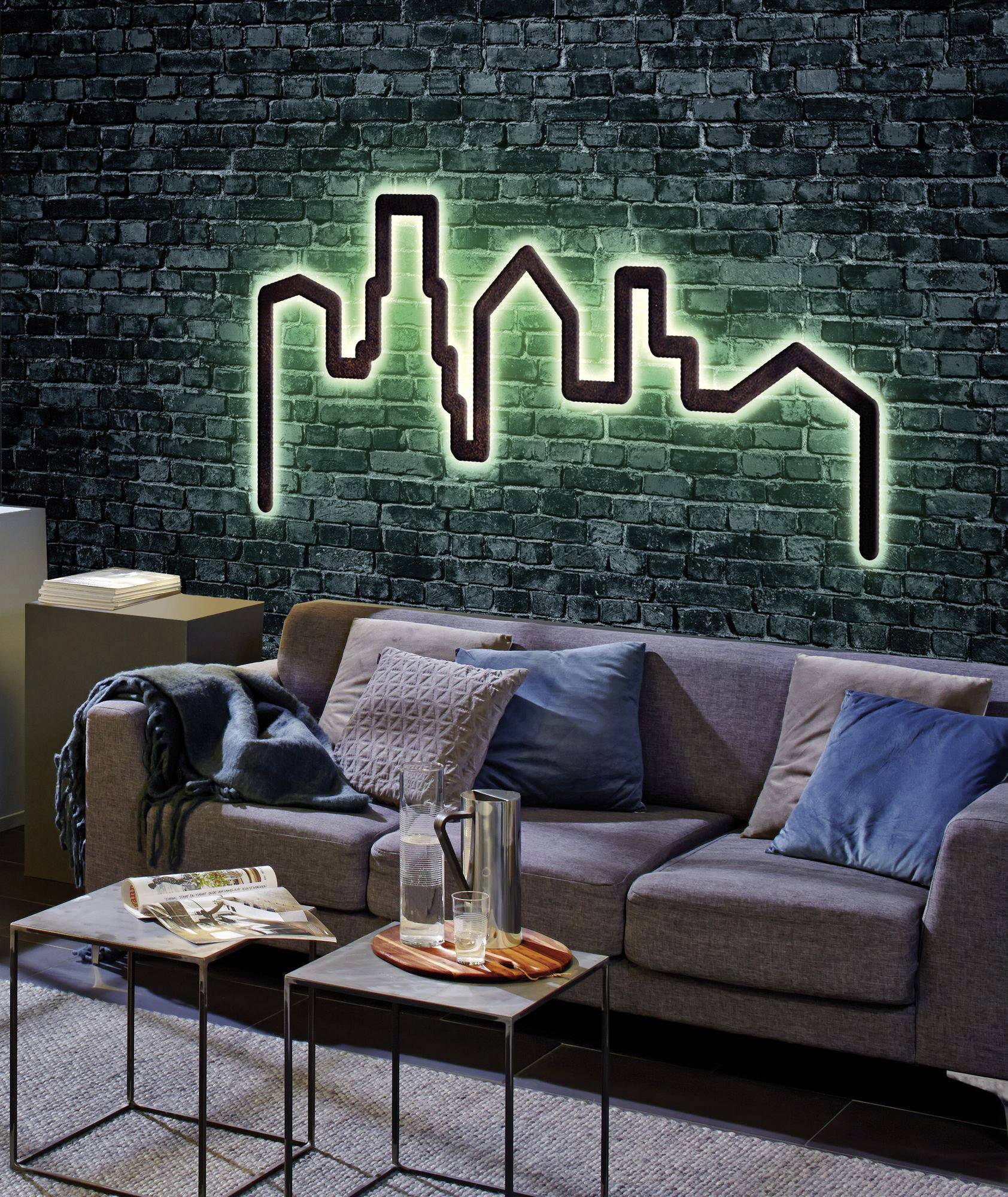 A modern living room with a grey sofa, blue cushions, a throw, and two small side tables. A luminous skyline decoration adorns the wall.