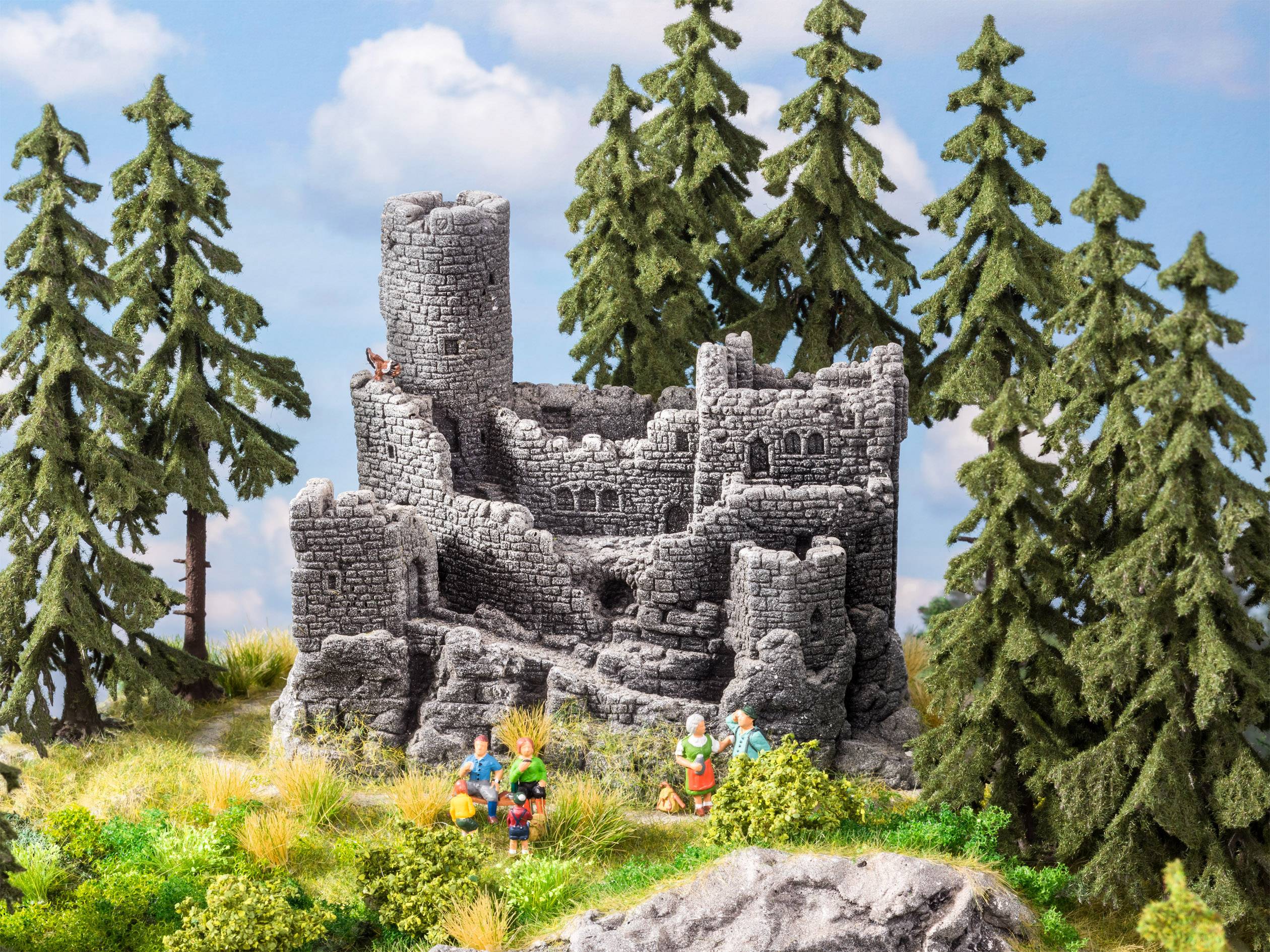A miniature landscape depicts a medieval castle ruin surrounded by tall pine trees. In the foreground, small figures can be seen.