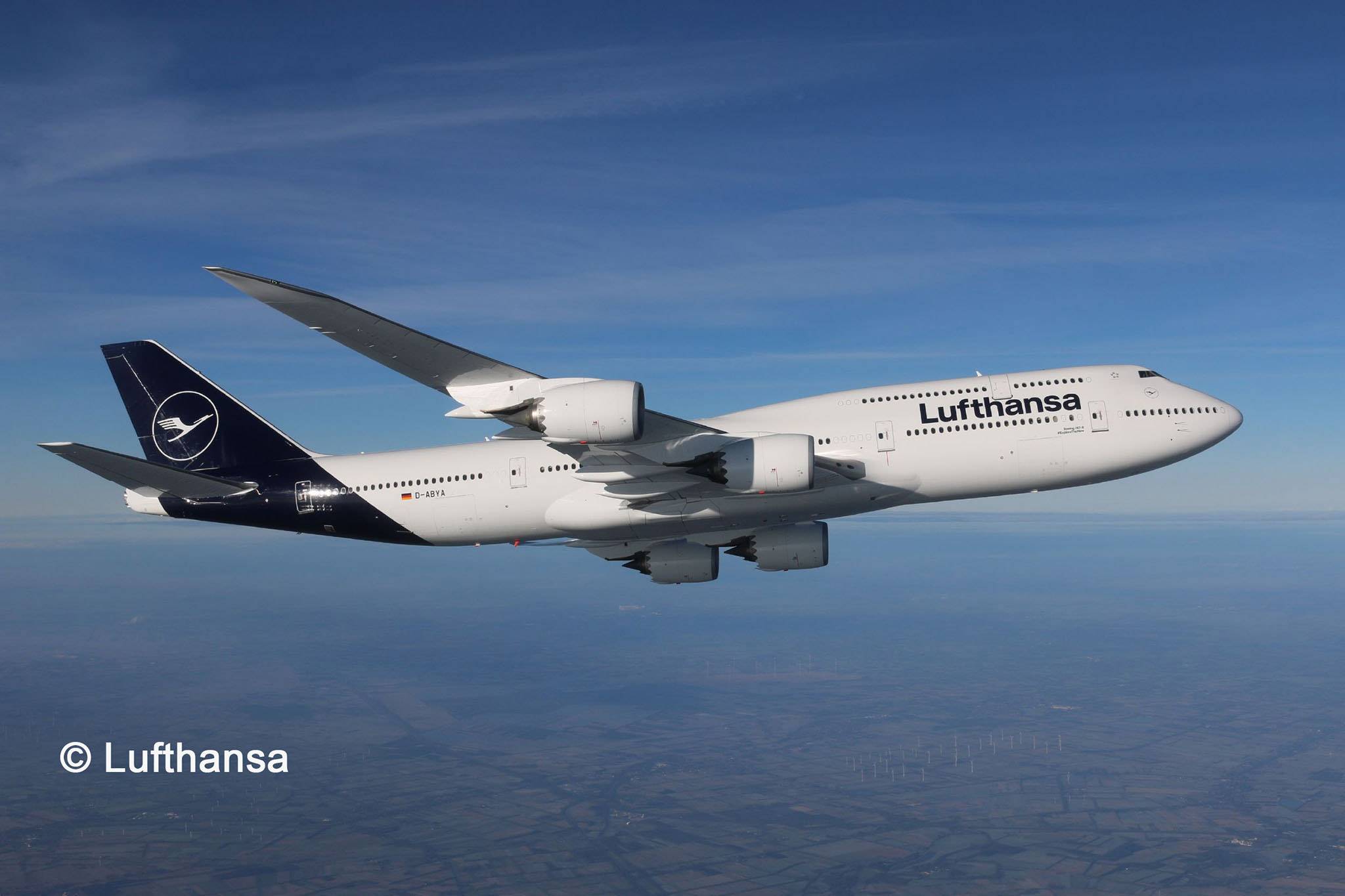 A large Lufthansa aircraft flies at a great altitude over a landscape, visible against a clear sky.