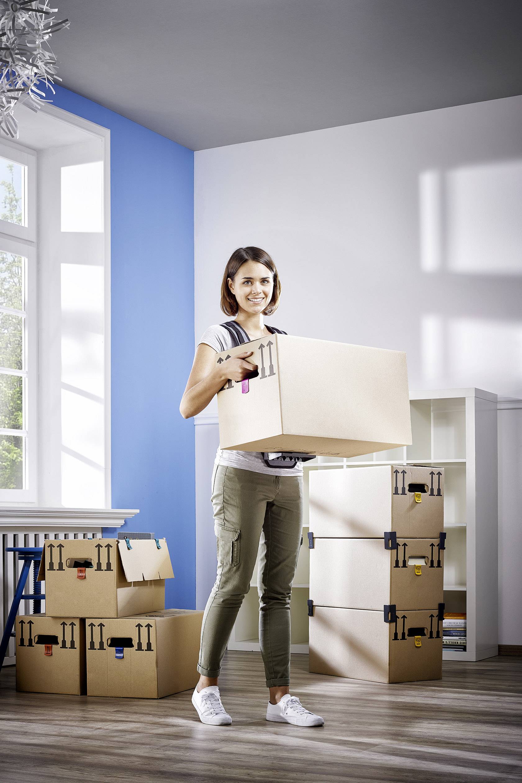 A person is standing smiling in a room, holding a removal box. More stacked boxes can be seen in the background.