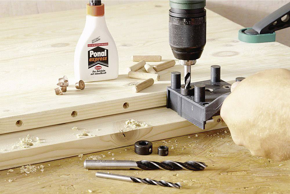 A hand is using a drilling template and a drill bit to bore holes in wood. Wooden dowels and wood glue are on the table.