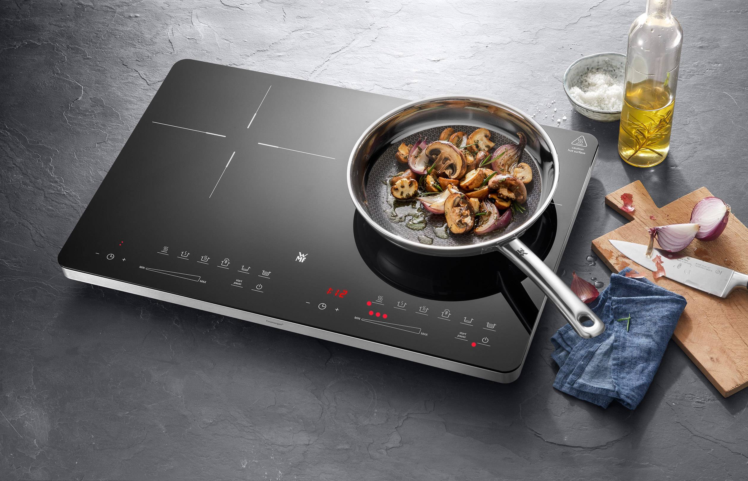 A modern induction hob with a pan of mushroom and onion dish. Beside it, a chopping board with sliced onions.