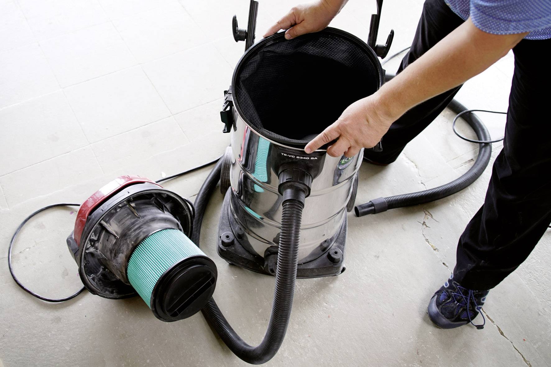A person opens a vacuum cleaner container to check or replace the filter. The vacuum cleaner is standing on a floor.