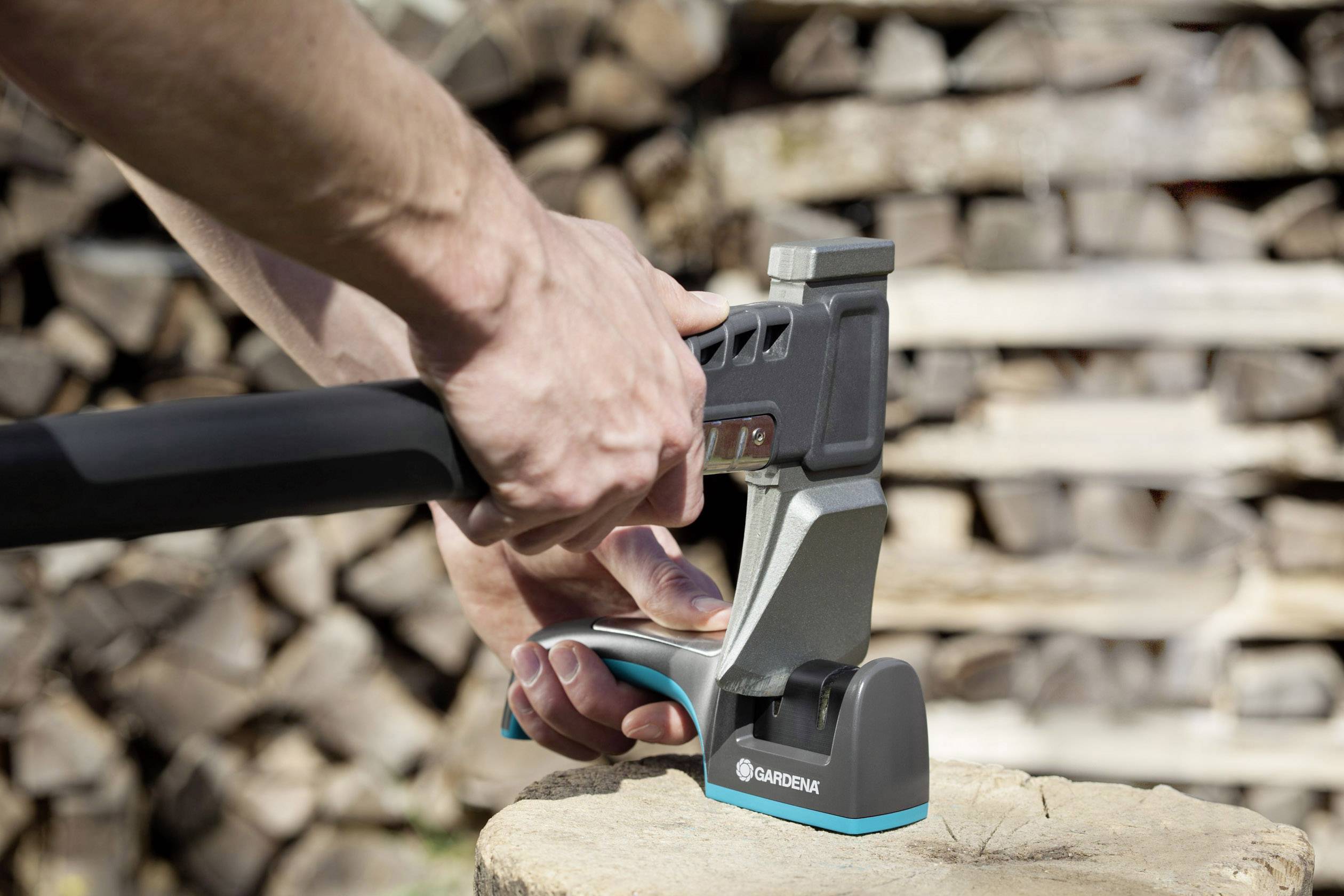 Two hands are holding an axe with a metal guard on a tree stump; stacked logs can be seen in the background.