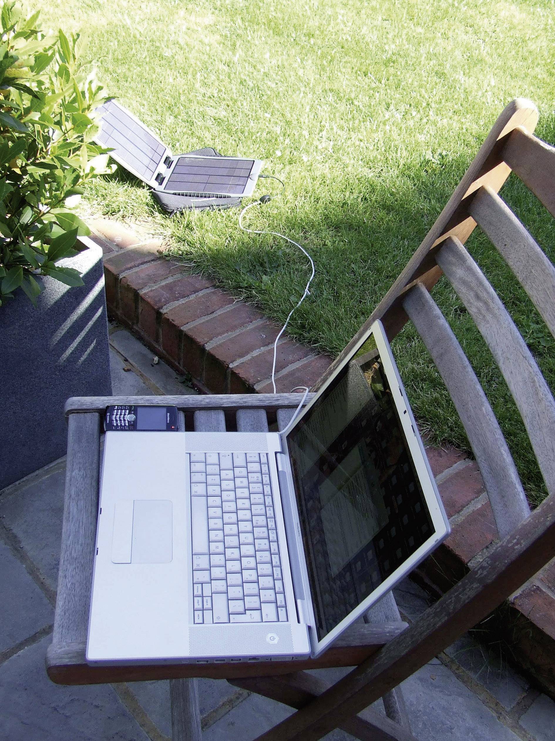 A laptop on a wooden chair outdoors, connected to a solar panel on the grass, demonstrates eco-friendly energy.