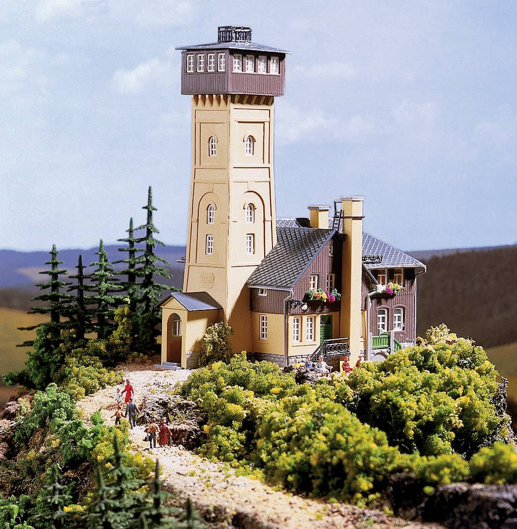 Model of a landscape with a tall tower and an adjacent building, surrounded by trees and small figures on a path.