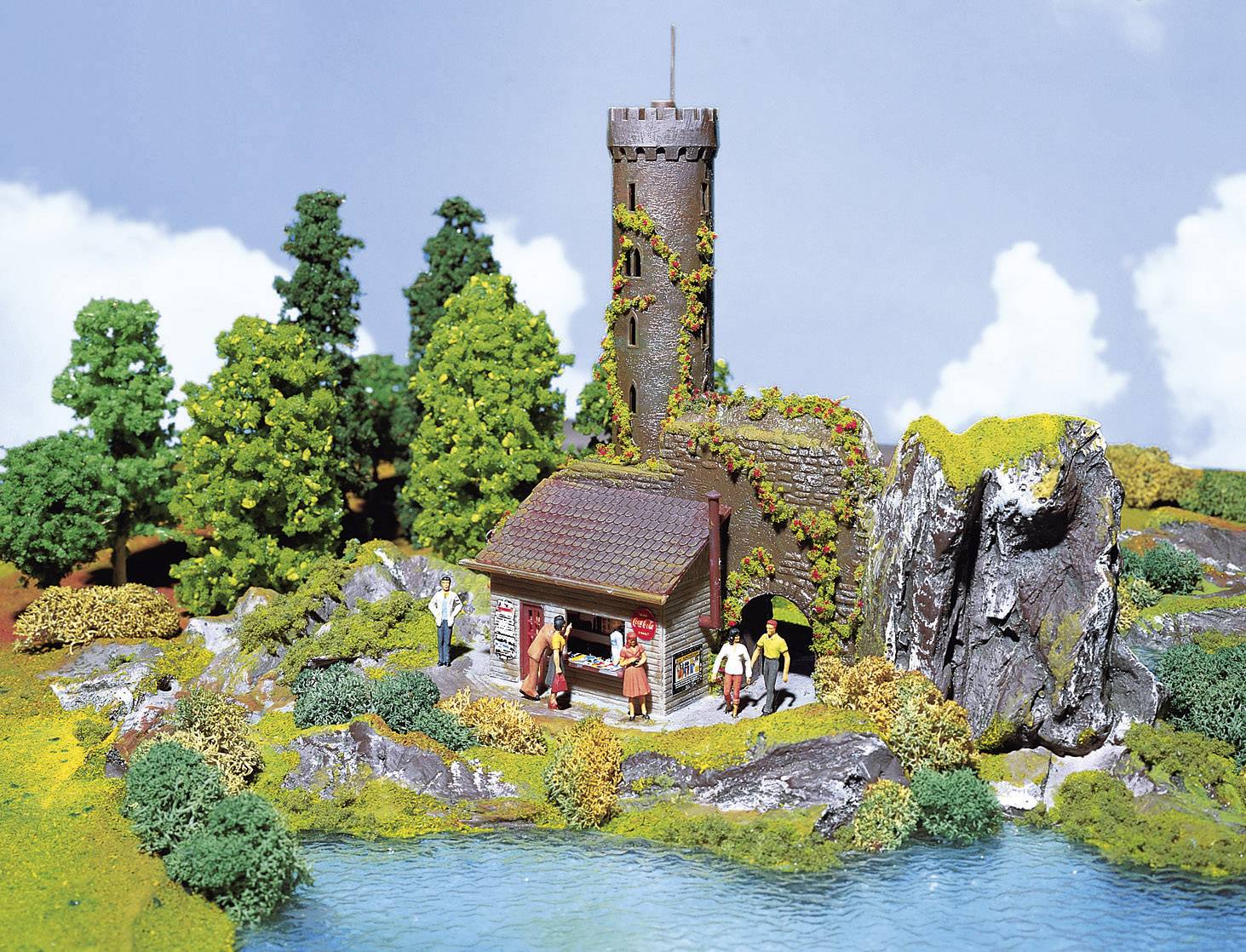 A miniature model depicts a medieval castle with a tower, surrounded by trees and a pond. Figurines bring the scene to life.