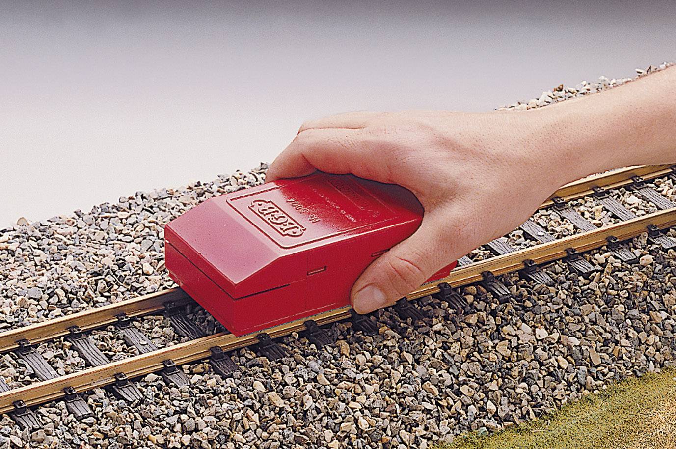 A hand holds a red rail cleaning device on a model railway track over ballast, which is used for cleaning the rails.