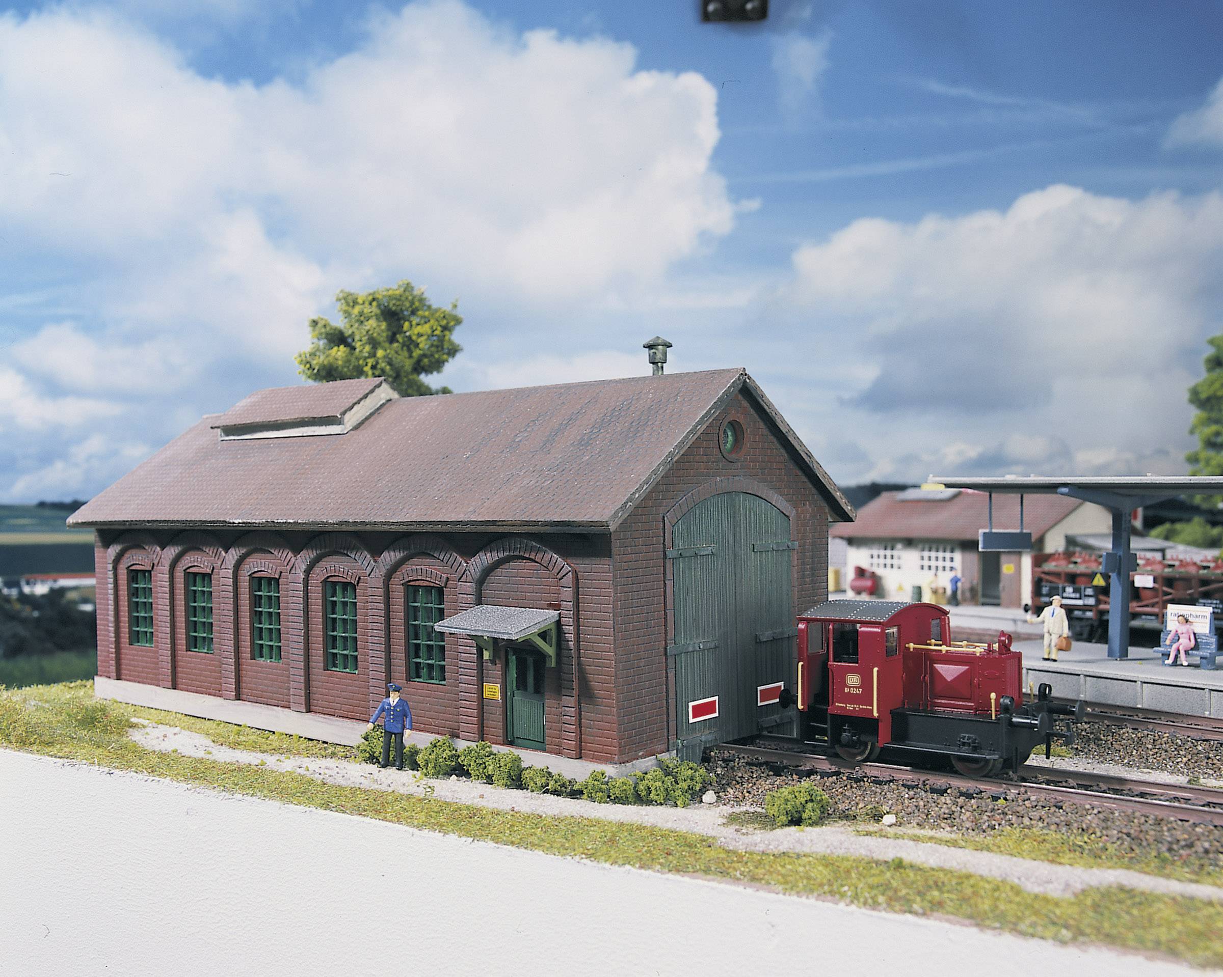A model of an old engine shed with a small red locomotive in front of it. People are standing nearby on a platform.