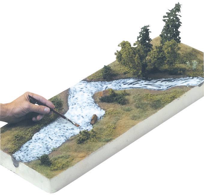 A hand is painting a river with a brush on a landscape diorama with trees, creating a lifelike model of a riverside scene.