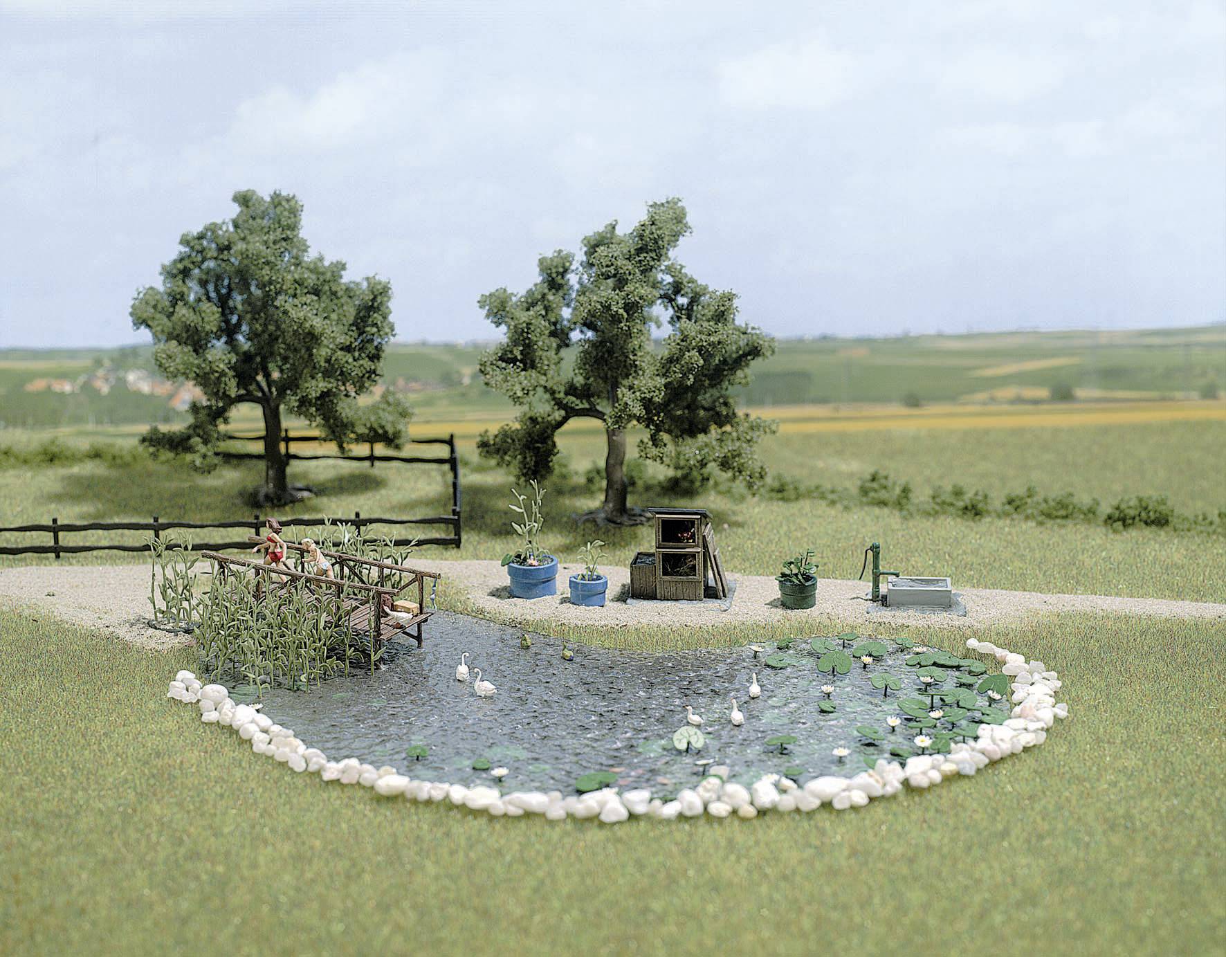 Miniature model of a rural landscape with a small pond, ducks, trees and a fence along the pond's edge. Meadows in the background.