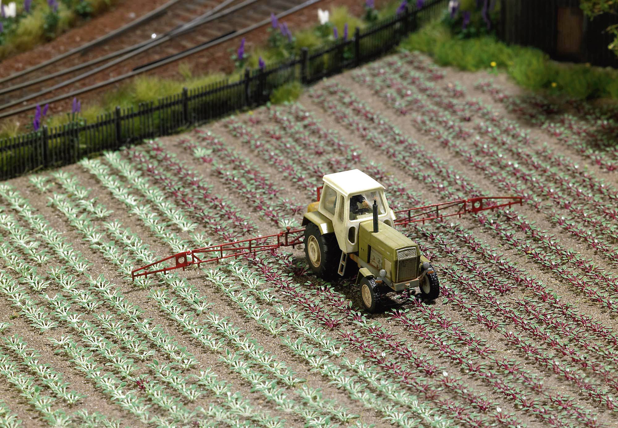 A small tractor drives across a miniature field and sprays pesticides on plants. A railway track runs in the background.